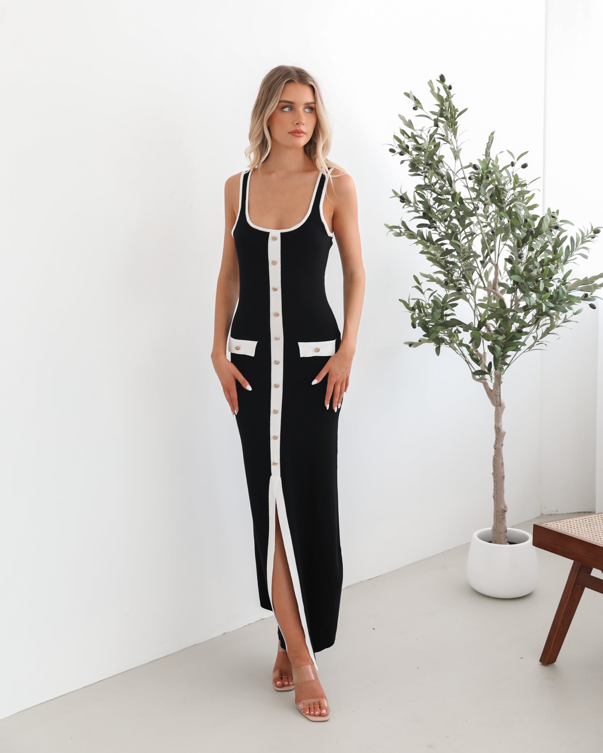 A model wearing a black sleeveless knit dress with a scoop neckline, center-front buttons, and a maxi length hem.