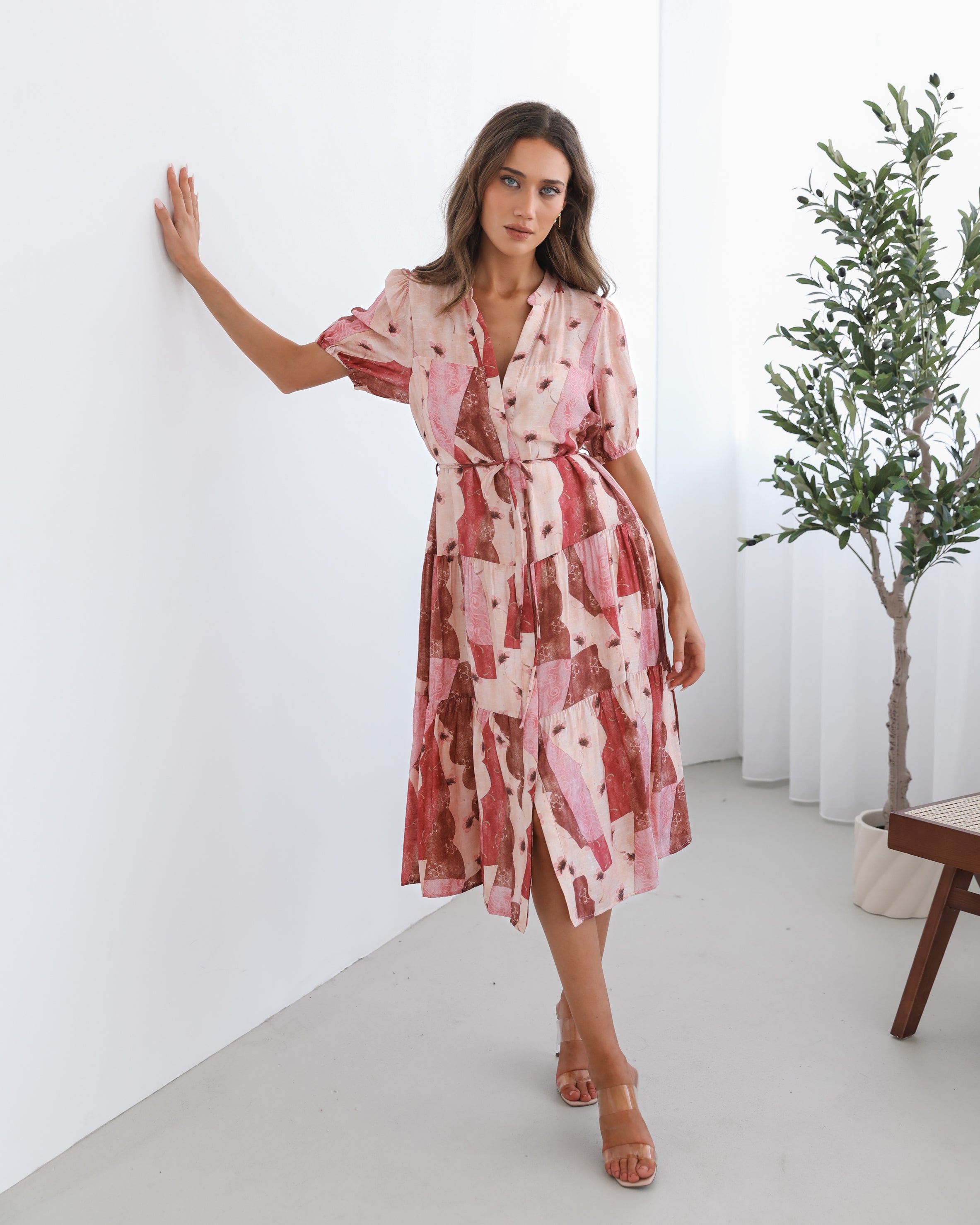 A woman standing in a room with a plant in the background, wearing a multi-toned print dress with short puff sleeves, a collar with a v-neck, center-front buttons, and a tiered skirt.