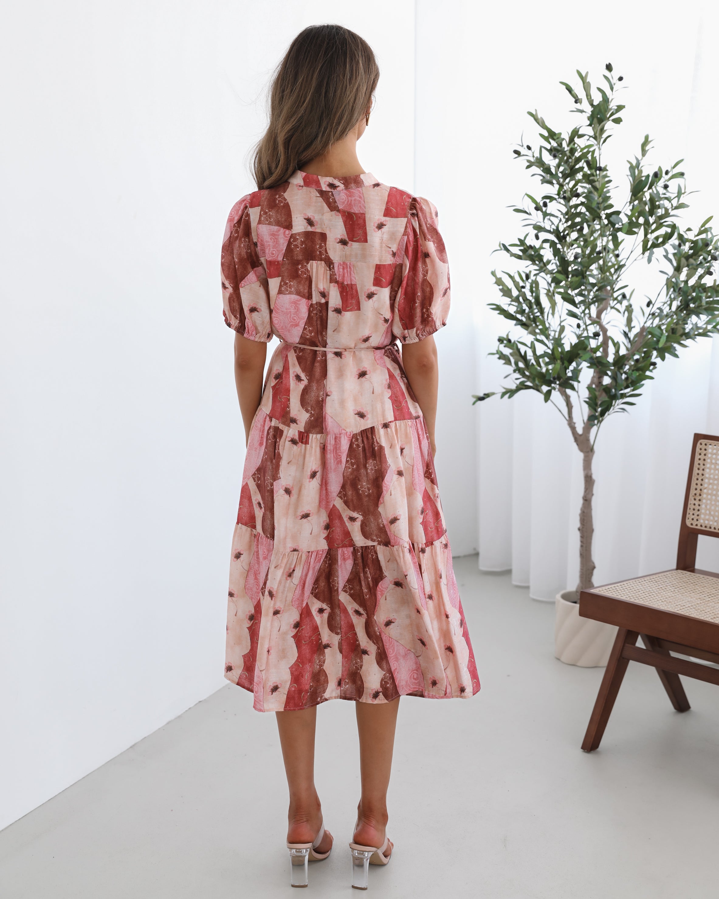 A woman standing in a room with a plant in the background, wearing a multi-toned print dress with short puff sleeves, a collar with a v-neck, center-front buttons, and a tiered skirt.