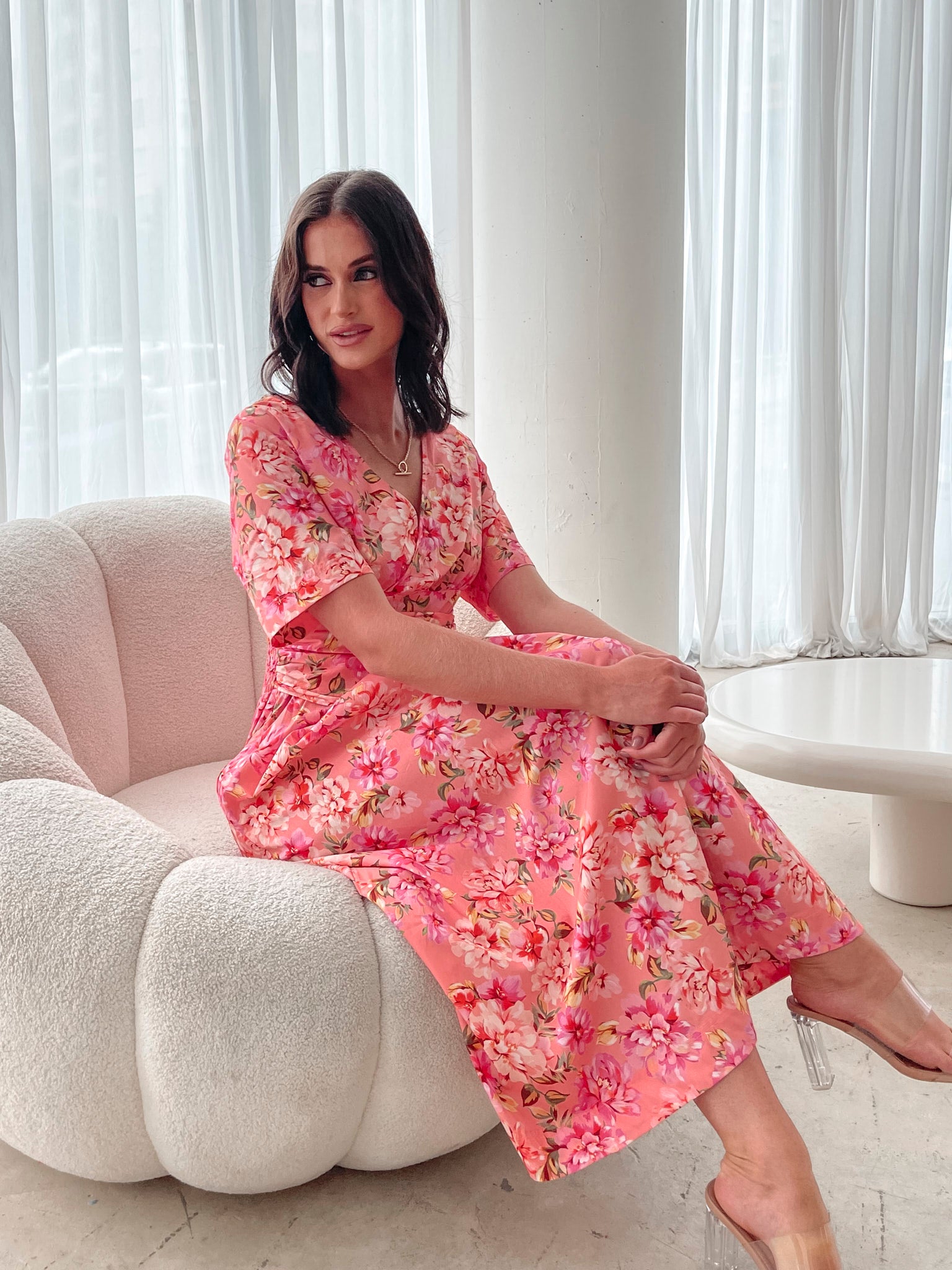 A person sitting on a white sofa wearing a peach blossom midi dress with a wide v-neckline, short puff sleeves, and a flowy a-line skirt.