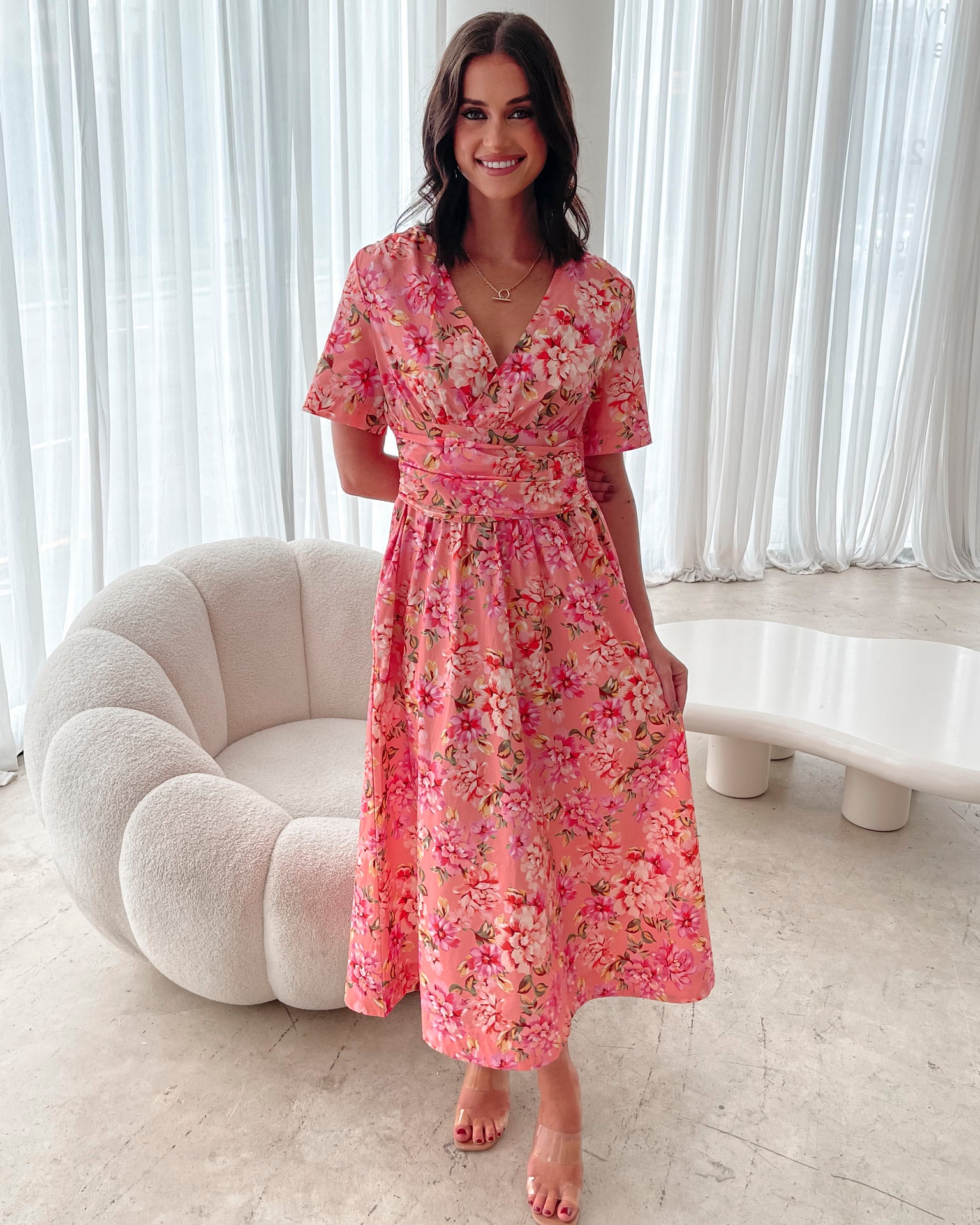 A person sitting on a white sofa wearing a peach blossom midi dress with a wide v-neckline, short puff sleeves, and a flowy a-line skirt.
