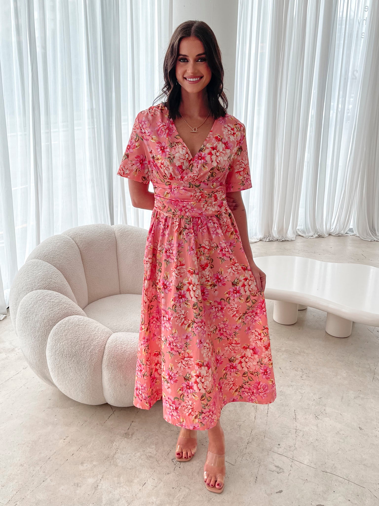A person sitting on a white sofa wearing a peach blossom midi dress with a wide v-neckline, short puff sleeves, and a flowy a-line skirt.