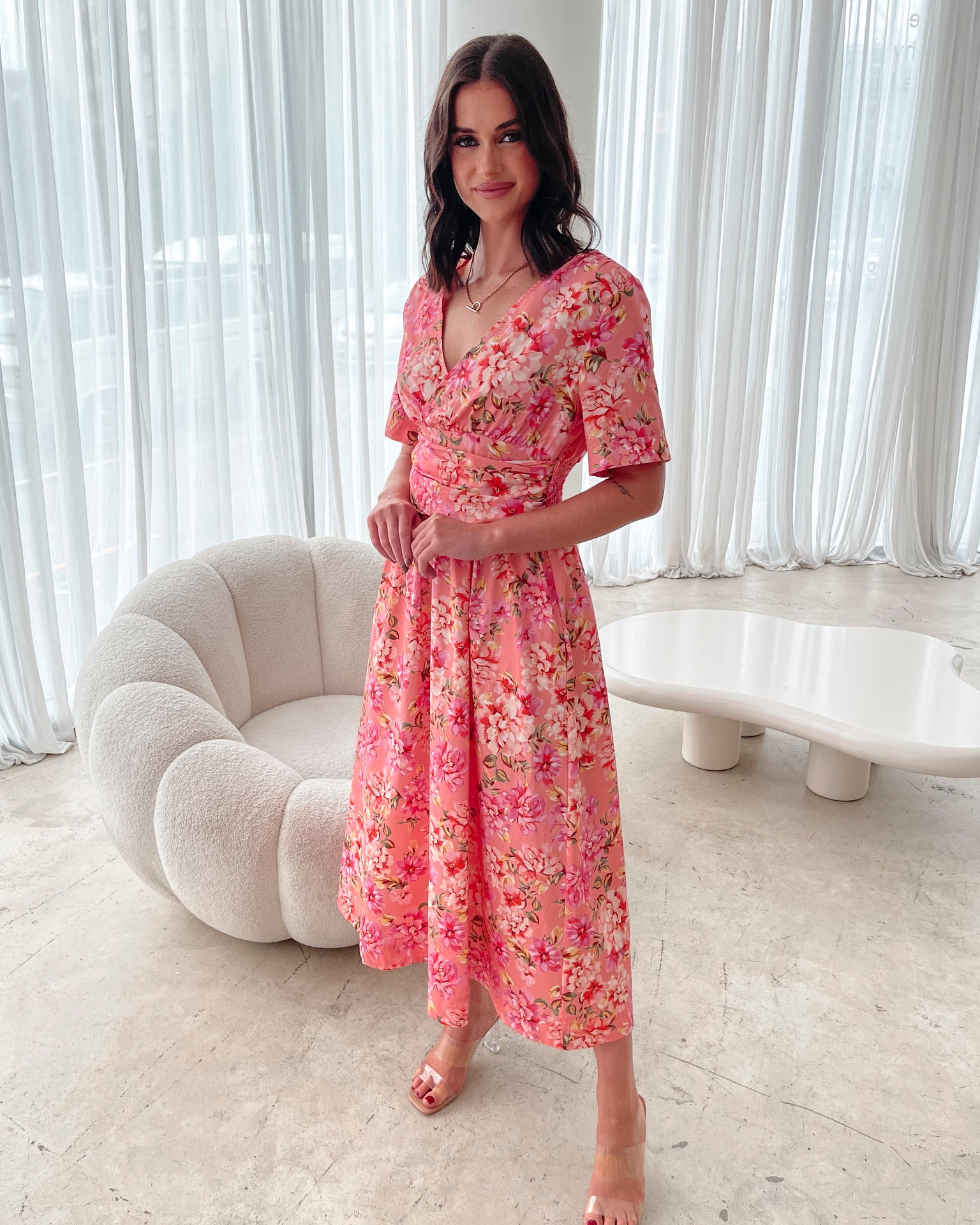 A person sitting on a white sofa wearing a peach blossom midi dress with a wide v-neckline, short puff sleeves, and a flowy a-line skirt.