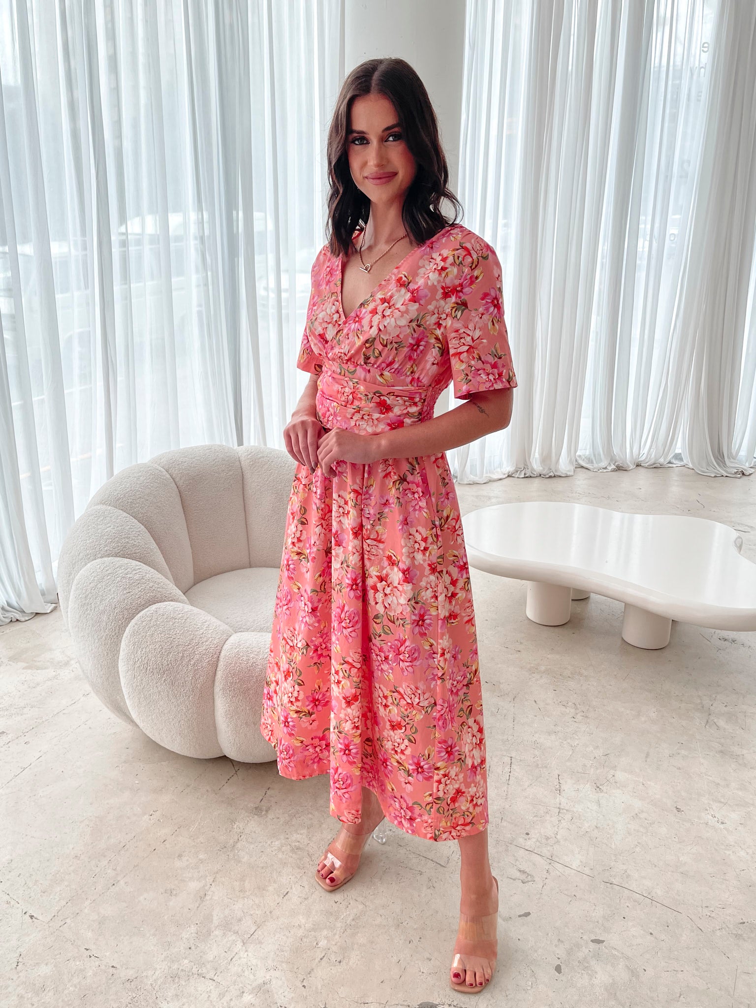A person sitting on a white sofa wearing a peach blossom midi dress with a wide v-neckline, short puff sleeves, and a flowy a-line skirt.