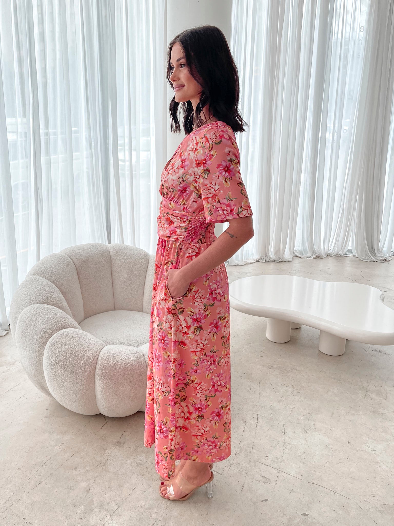 A person sitting on a white sofa wearing a peach blossom midi dress with a wide v-neckline, short puff sleeves, and a flowy a-line skirt.