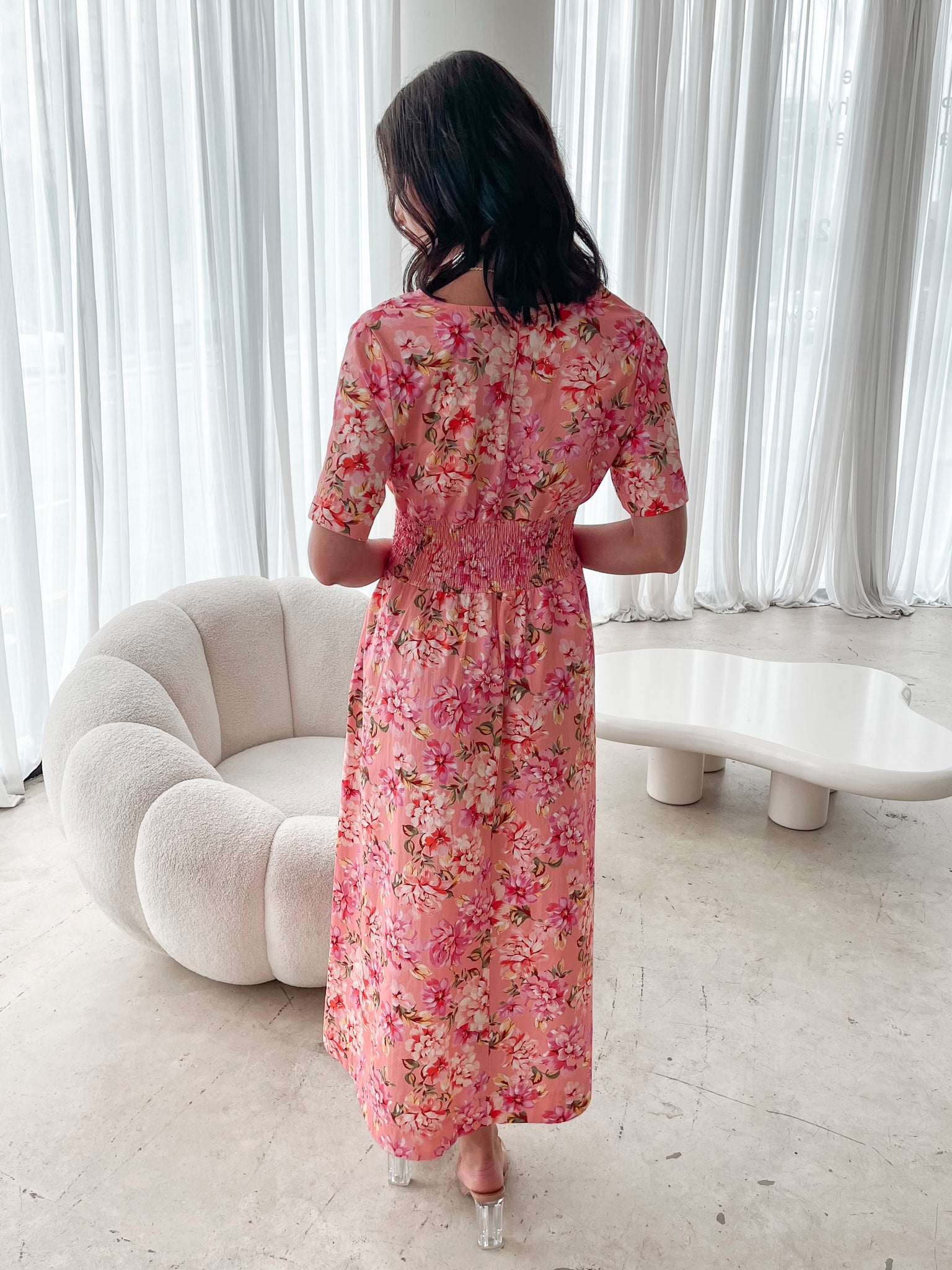 A person sitting on a white sofa wearing a peach blossom midi dress with a wide v-neckline, short puff sleeves, and a flowy a-line skirt.