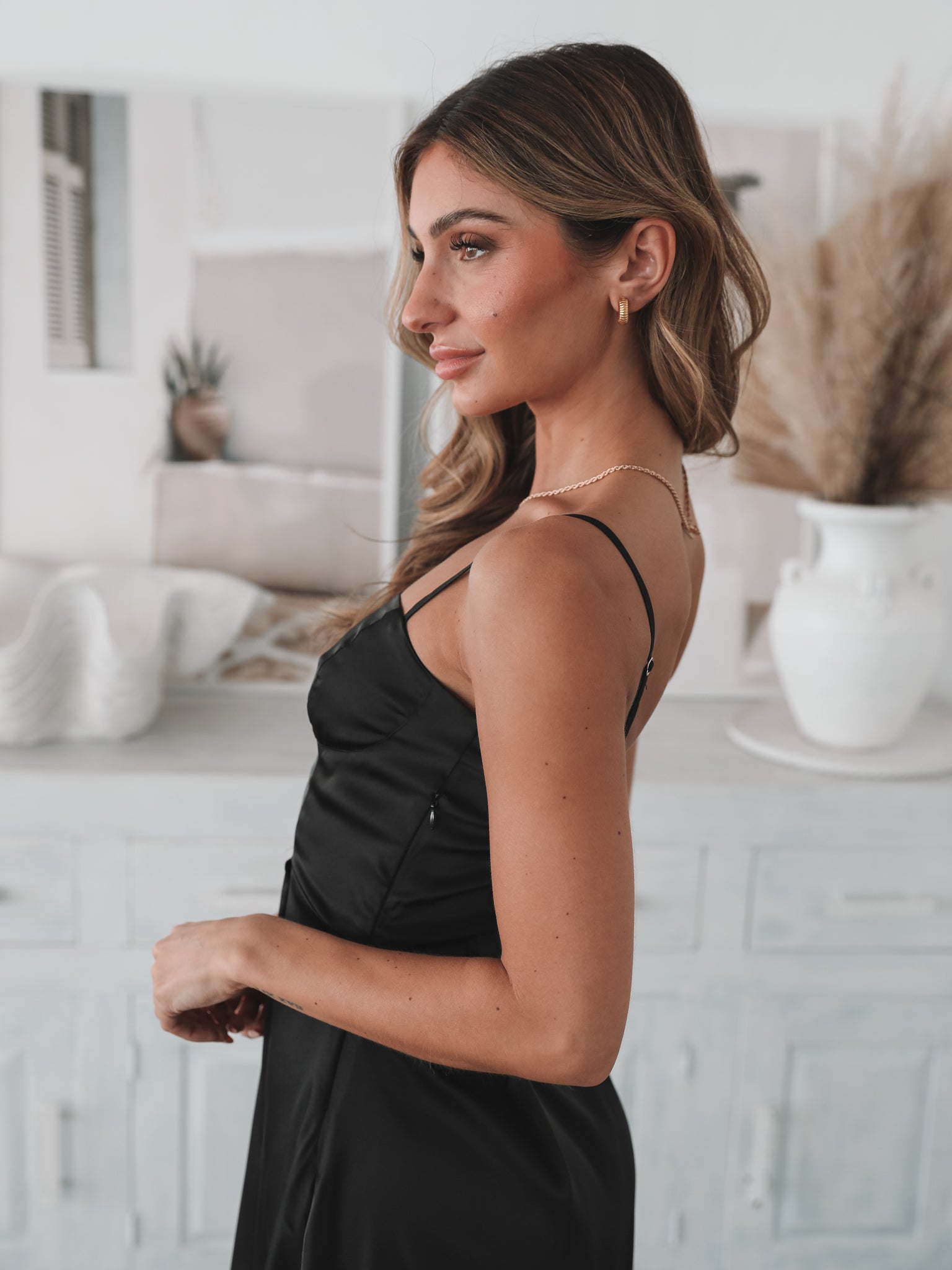 A woman standing in a room with light decor, wearing a black mini dress with a sweetheart neckline and slim straps. She has accessorized with a flower-shaped charm bracelet and is wearing block heels.