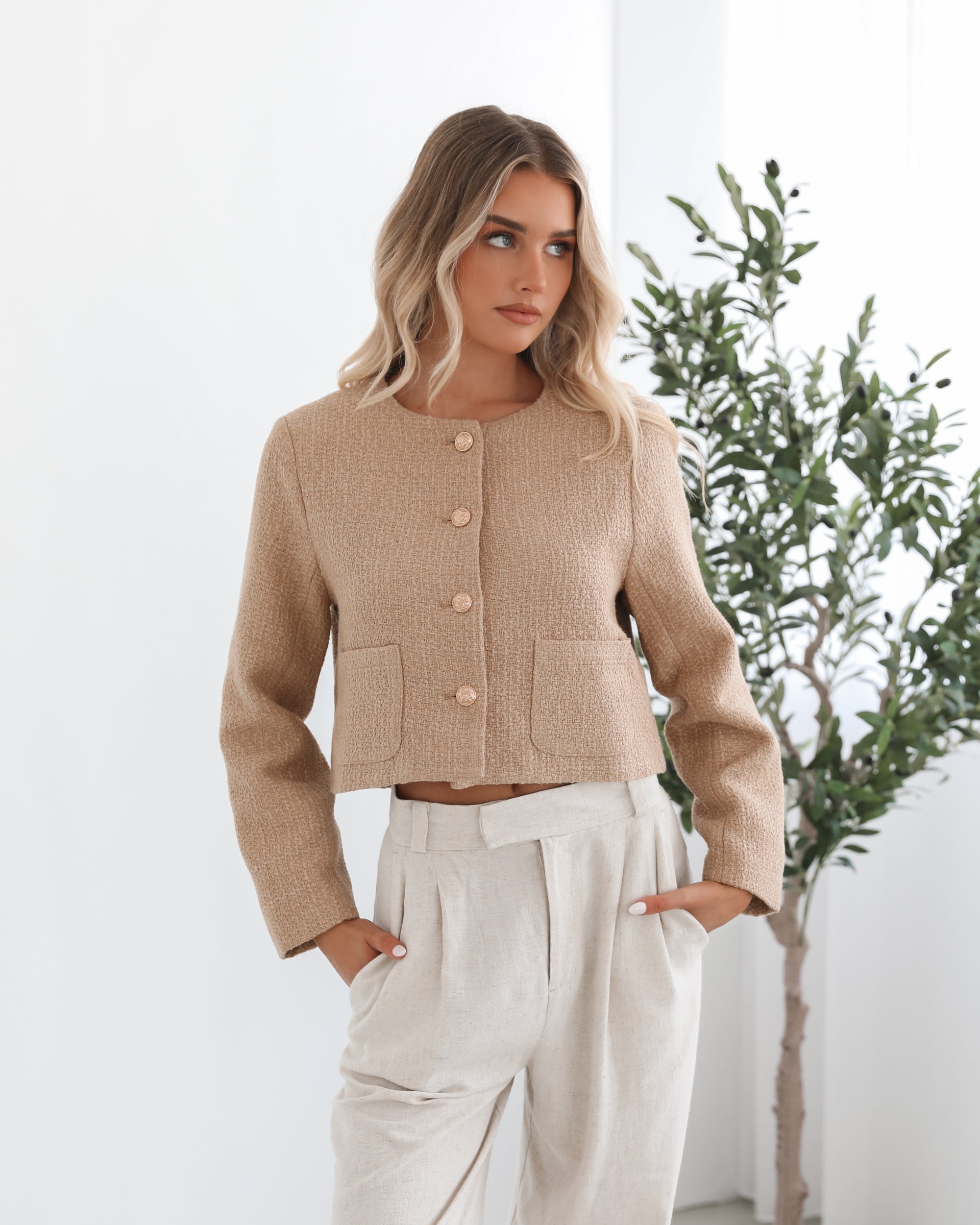 A woman wearing a beige Jacket with long sleeves, round neckline, and center-front buttons. The jacket has a crop length and she is paired with light-colored trousers.