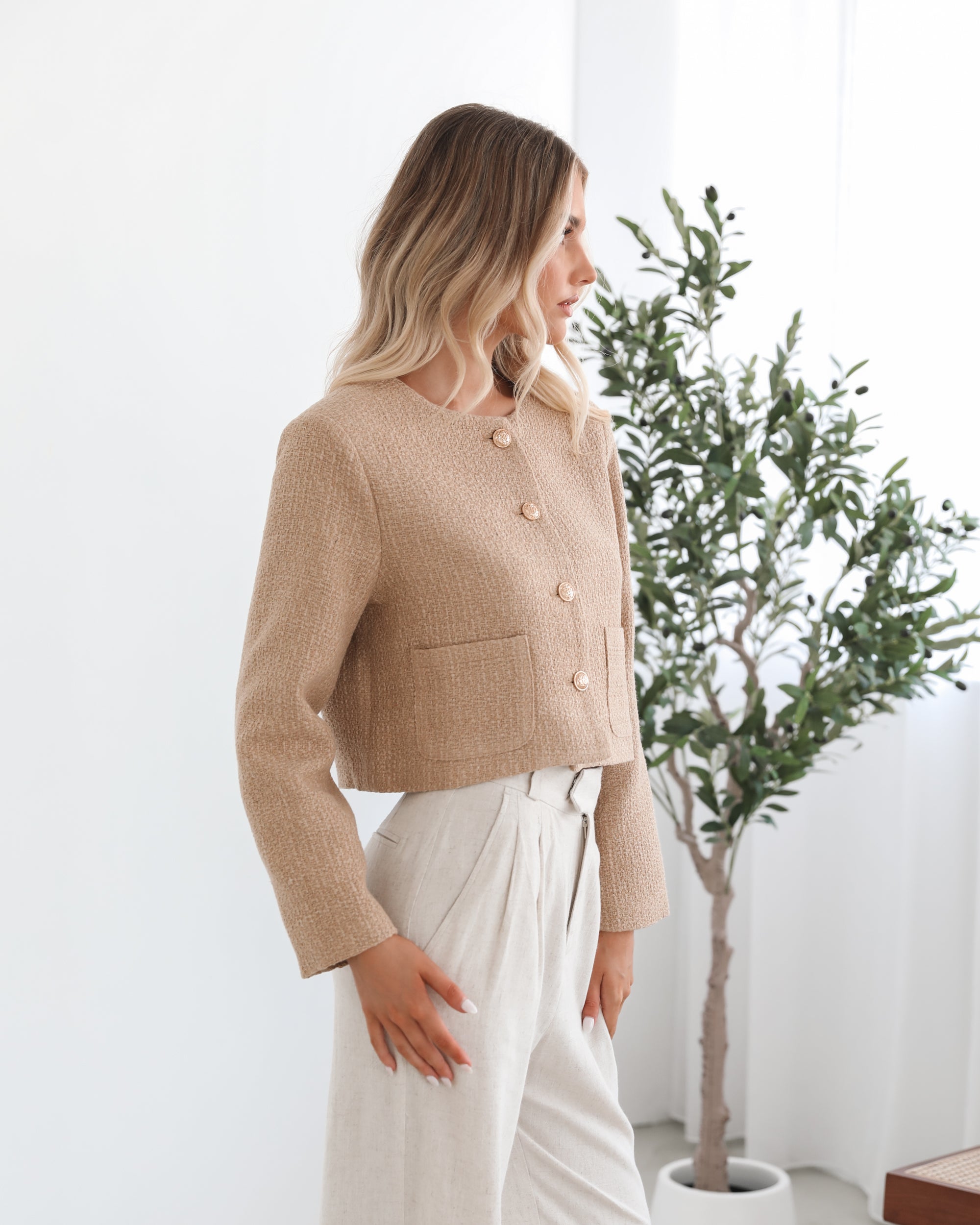 A woman wearing a beige Jacket with long sleeves, round neckline, and center-front buttons. The jacket has a crop length and she is paired with light-colored trousers.