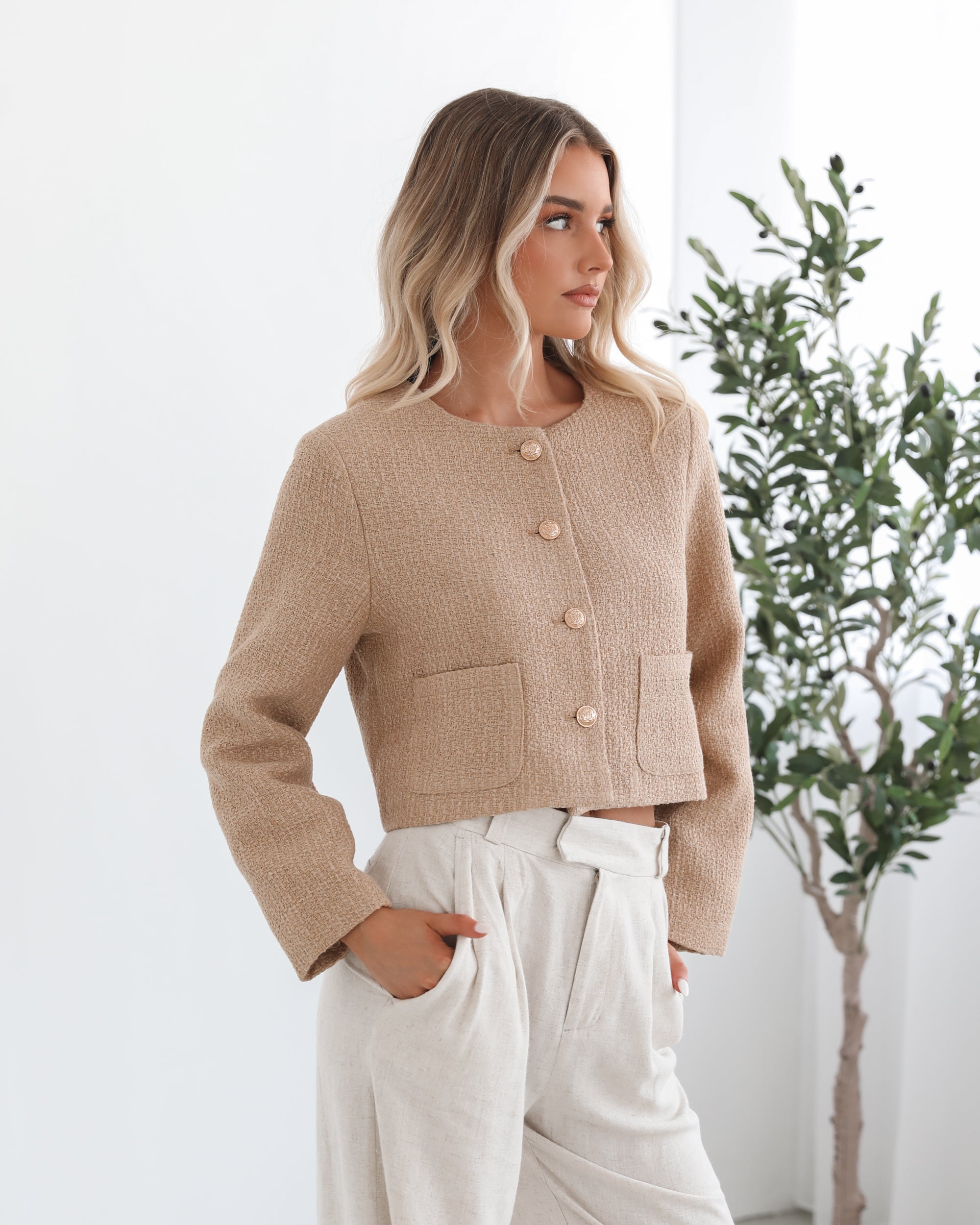 A woman wearing a beige Jacket with long sleeves, round neckline, and center-front buttons. The jacket has a crop length and she is paired with light-colored trousers.