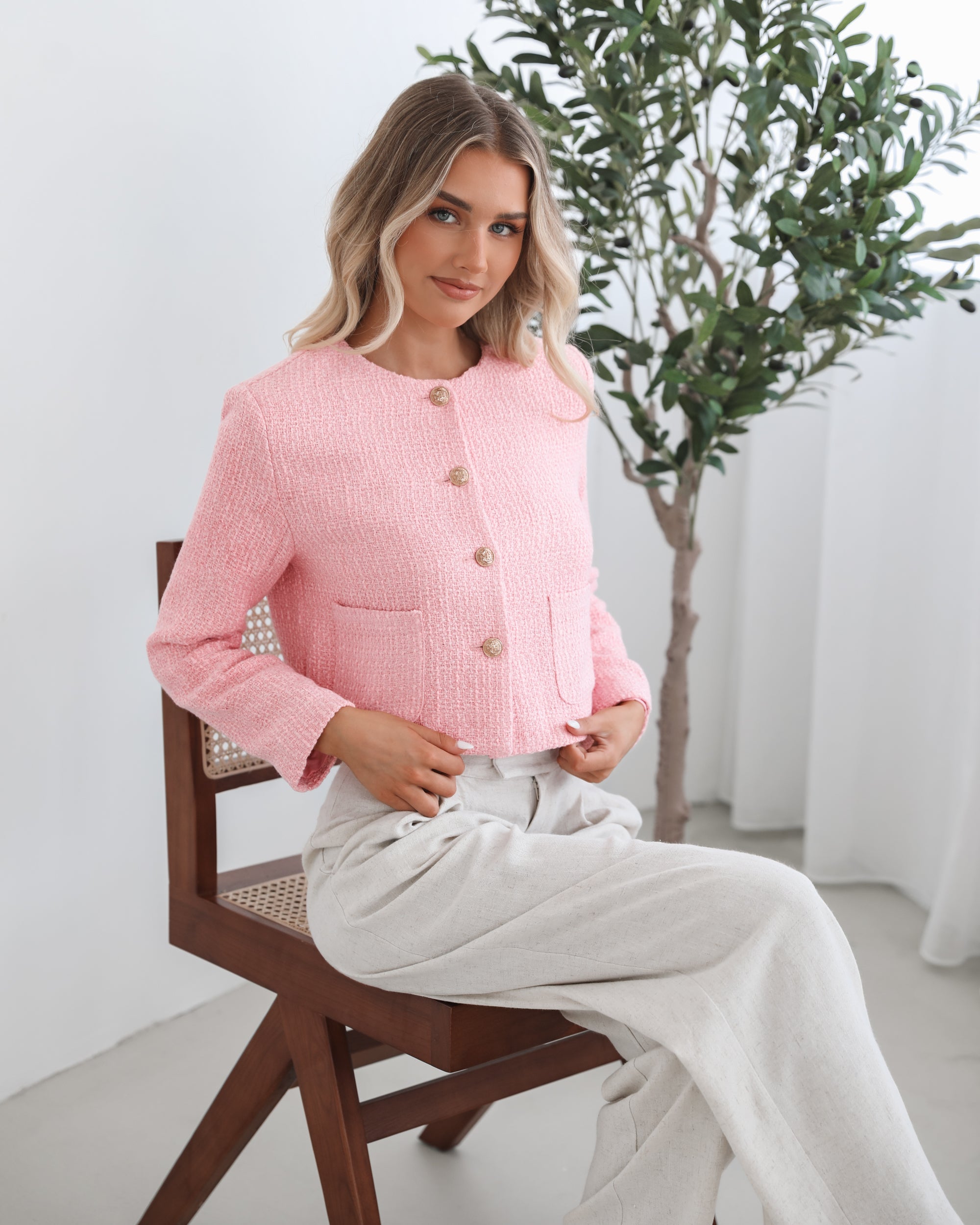 A woman wearing a pink Valdivia Jacket with long sleeves, round neckline, and center-front buttons. The jacket has a crop length and she is paired with light-colored trousers.