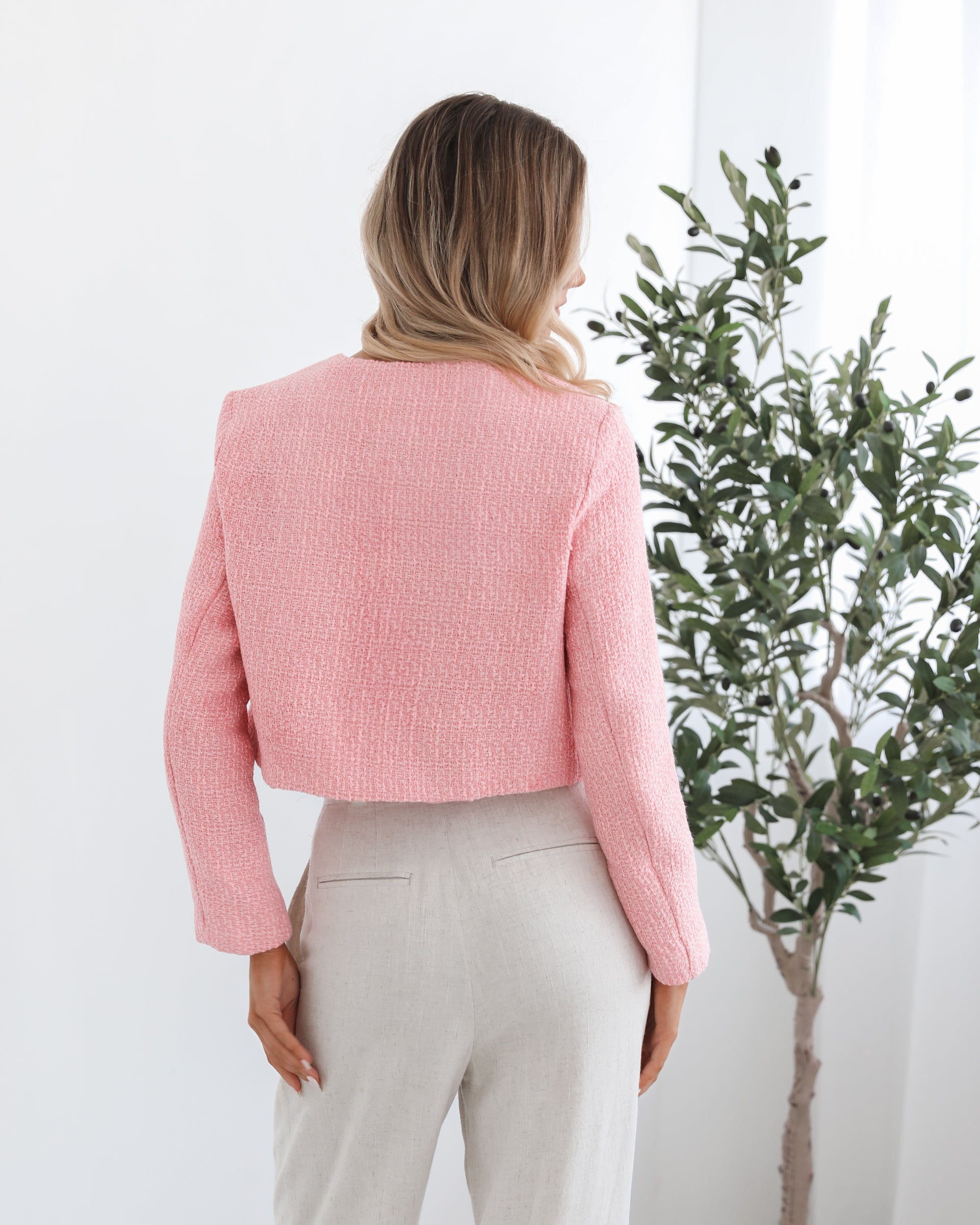 A woman wearing a pink Valdivia Jacket with long sleeves, round neckline, and center-front buttons. The jacket has a crop length and she is paired with light-colored trousers.