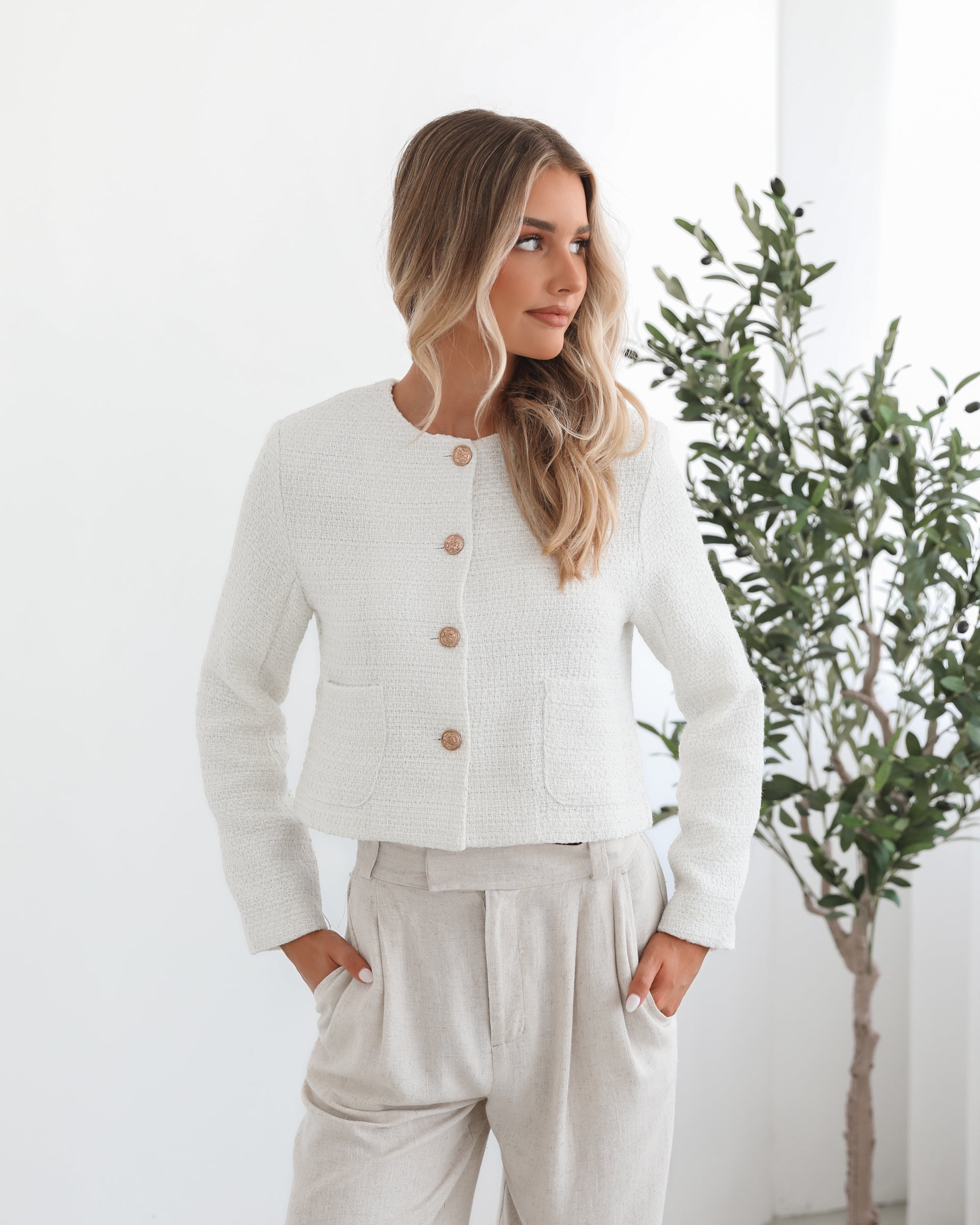 A woman sitting in a chair wearing a white Valdivia Jacket with long sleeves, round neckline, and center-front buttons. The jacket has a crop length and she is paired with a light-colored pants.