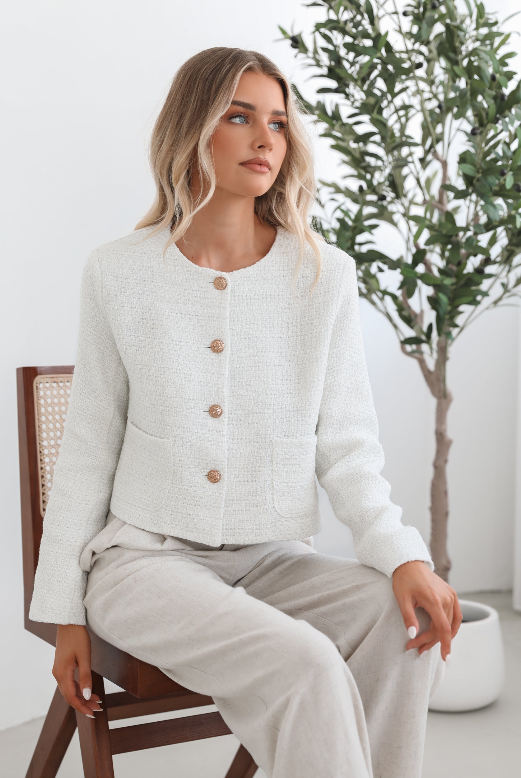 A woman sitting in a chair wearing a white Valdivia Jacket with long sleeves, round neckline, and center-front buttons. The jacket has a crop length and she is paired with a light-colored pants.