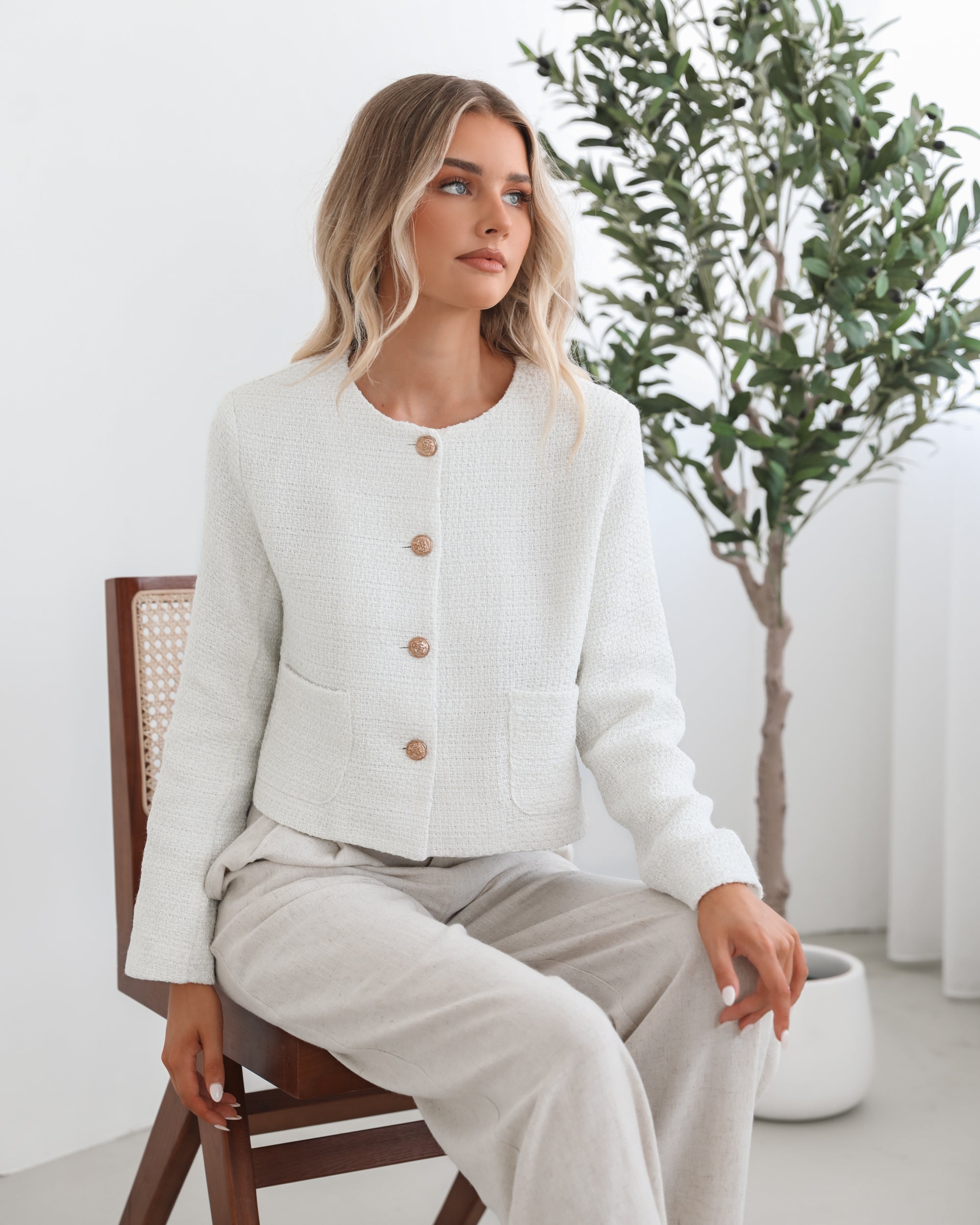 A woman sitting in a chair wearing a white Valdivia Jacket with long sleeves, round neckline, and center-front buttons. The jacket has a crop length and she is paired with a light-colored pants.