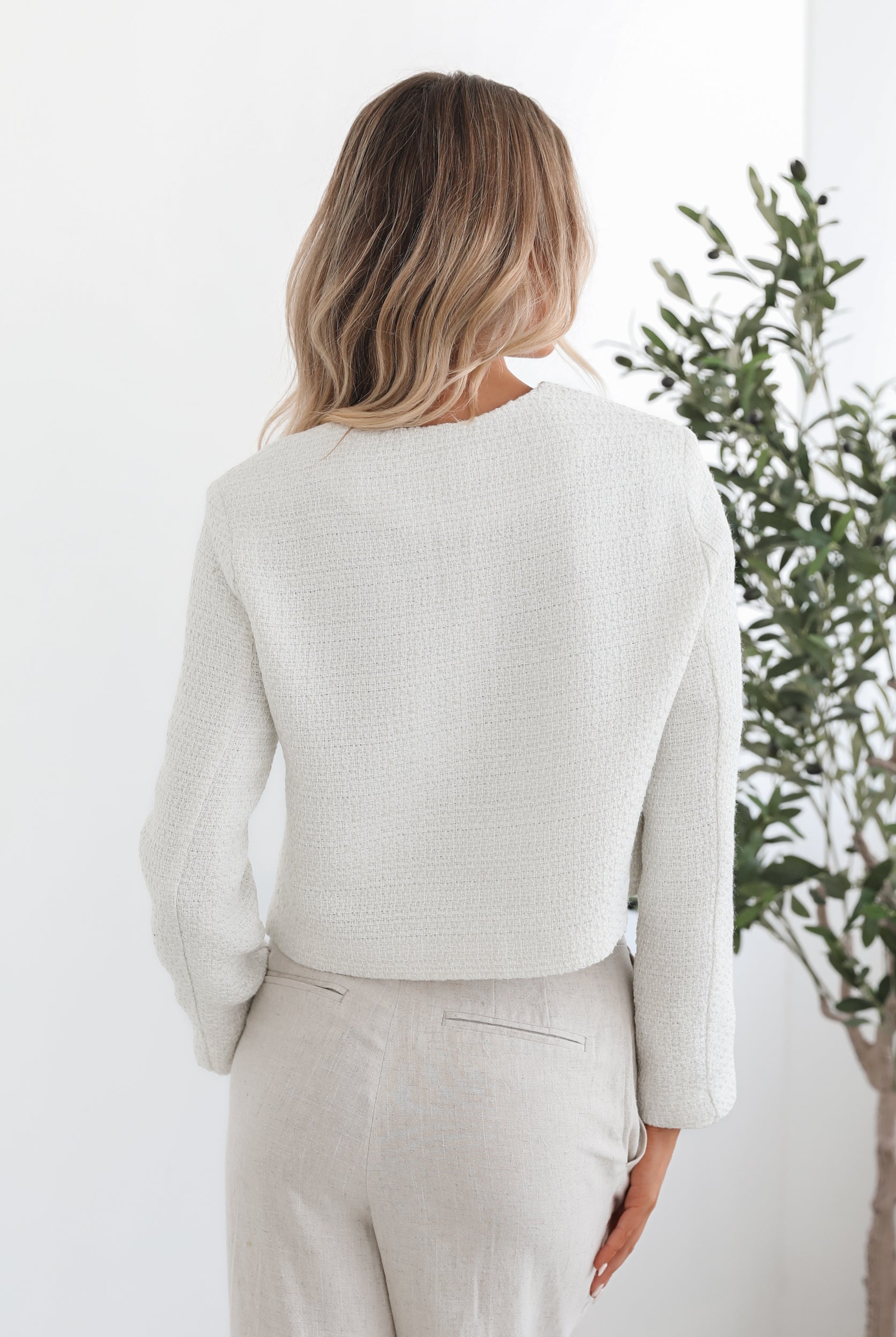 A woman sitting in a chair wearing a white Valdivia Jacket with long sleeves, round neckline, and center-front buttons. The jacket has a crop length and she is paired with a light-colored pants.