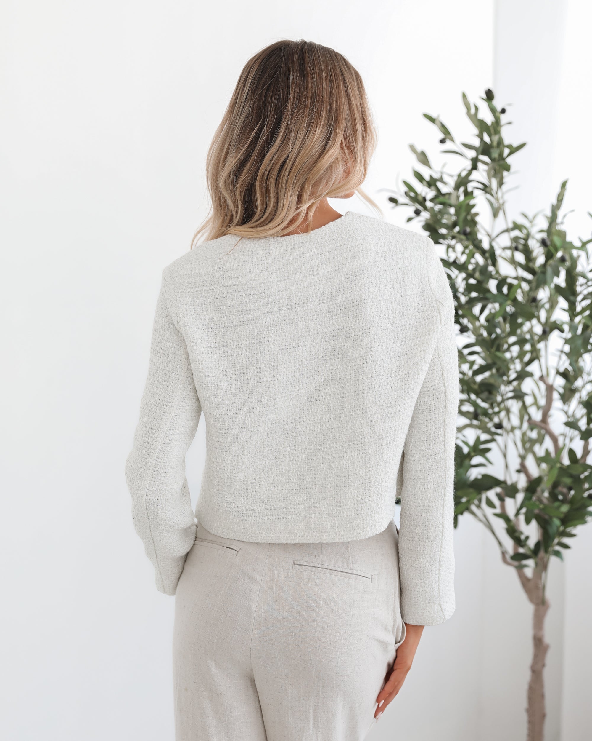A woman sitting in a chair wearing a white Valdivia Jacket with long sleeves, round neckline, and center-front buttons. The jacket has a crop length and she is paired with a light-colored pants.