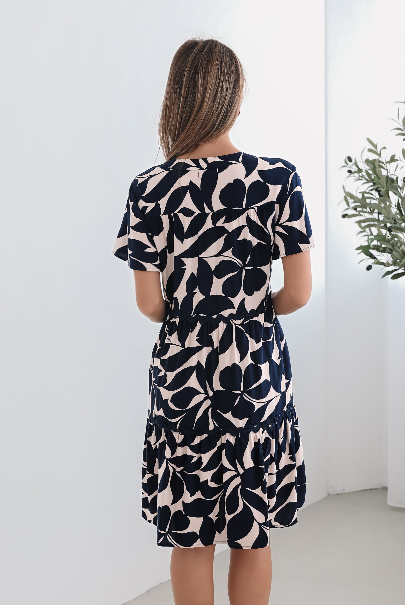 A woman standing in front of a plant, wearing a beige dress with navy-tone leaf prints, short flutter sleeves, and a v-neckline.