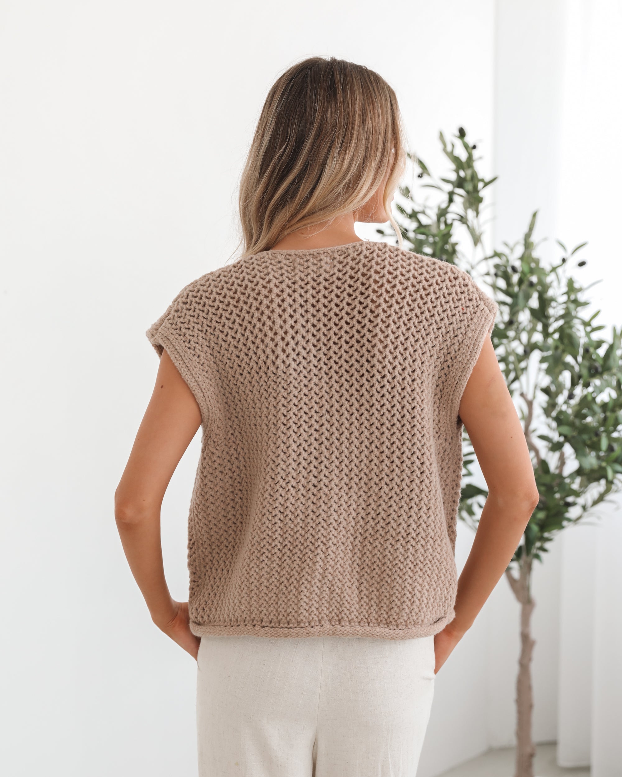 A woman wearing a beige knit top with round neckline, centre-front buttons, and a relaxed fit. The top is sleeveless and she is pairing it with light-colored pants.