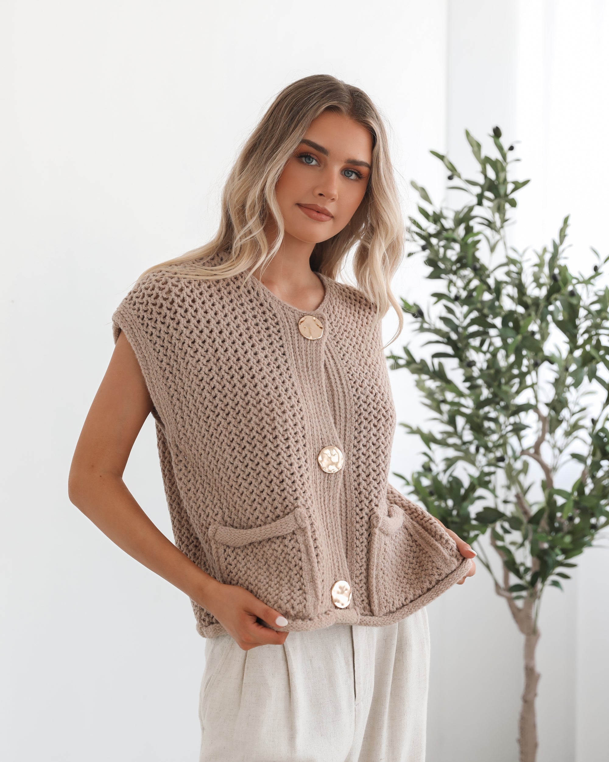 A woman wearing a beige knit top with round neckline, centre-front buttons, and a relaxed fit. The top is sleeveless and she is pairing it with light-colored pants.