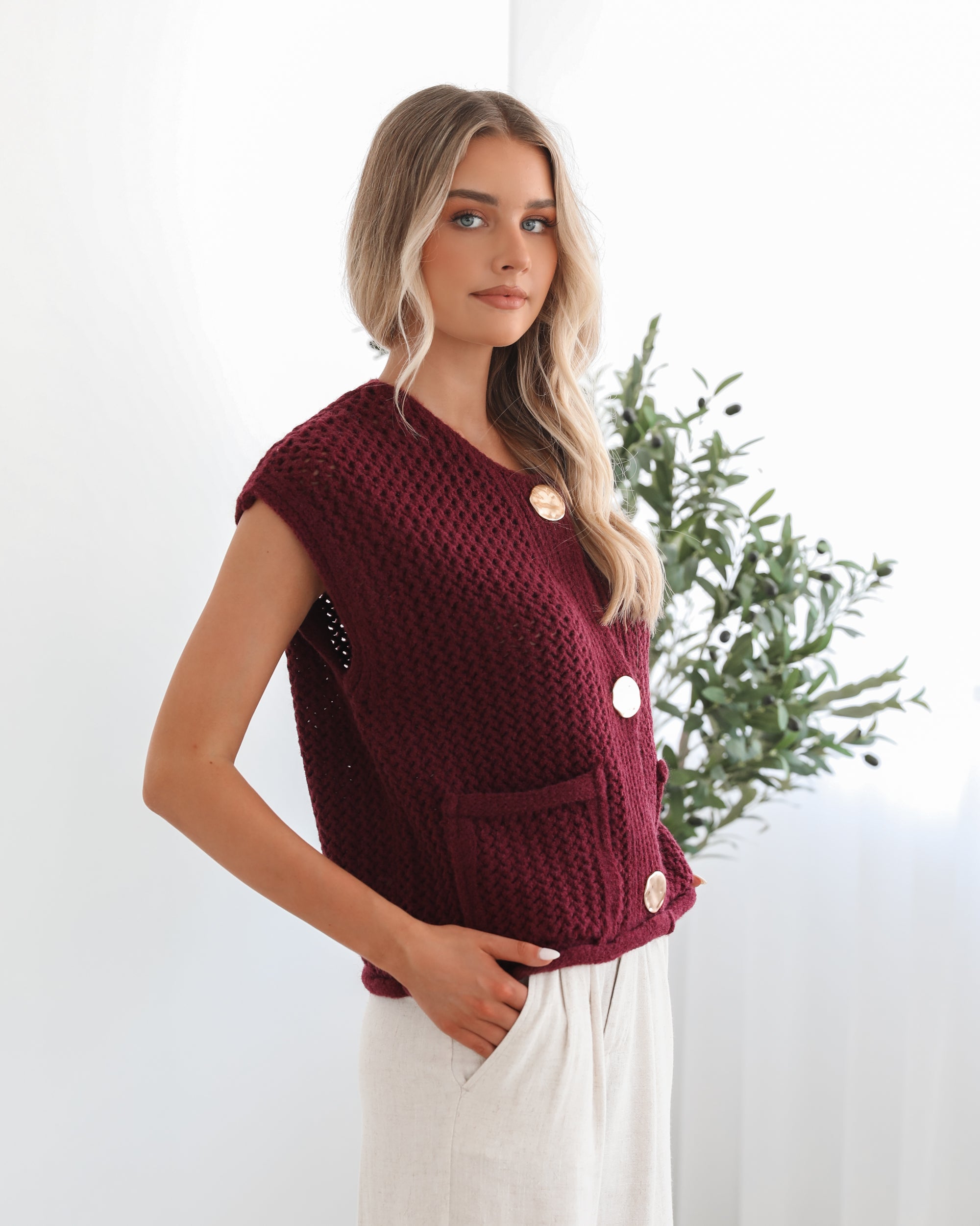 A woman wearing a sleeveless wine-colored knit top with round neckline and center-front buttons, paired with light-colored pants.