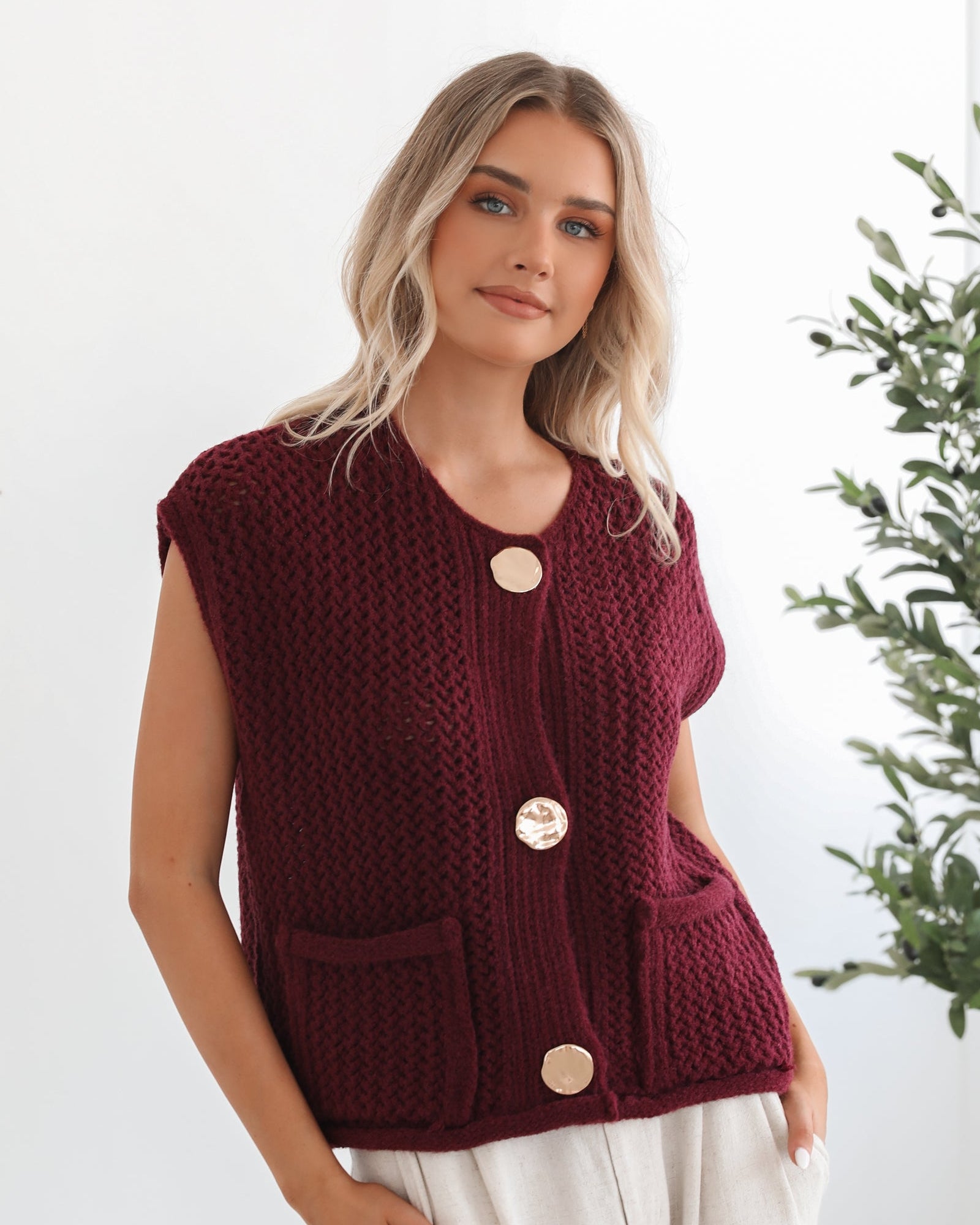 A woman wearing a sleeveless wine-colored knit top with round neckline and center-front buttons, paired with light-colored pants.
