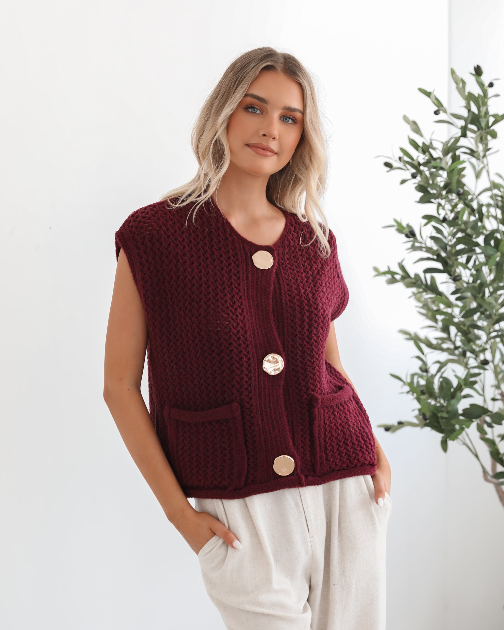 A woman wearing a sleeveless wine-colored knit top with round neckline and center-front buttons, paired with light-colored pants.