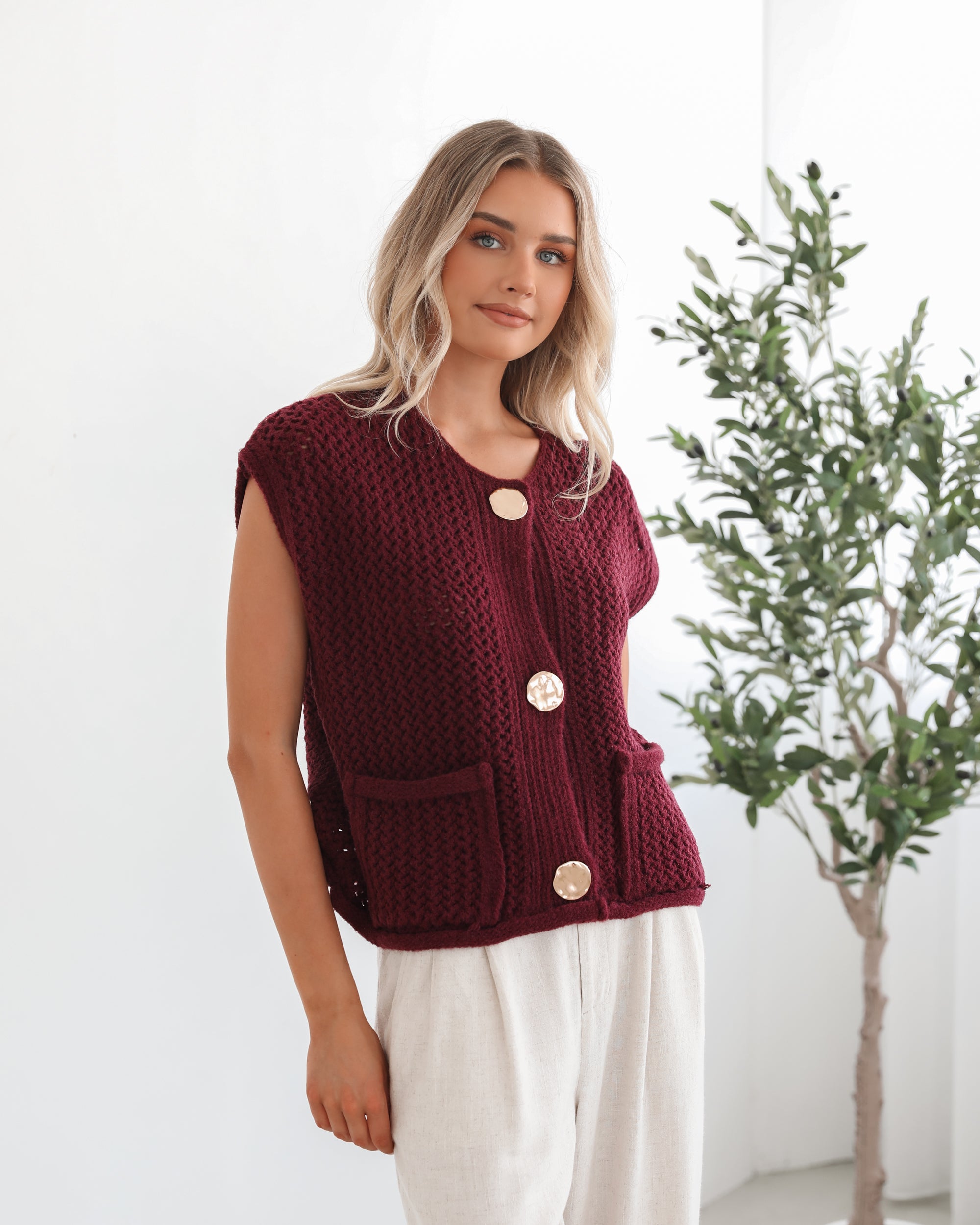 A woman wearing a sleeveless wine-colored knit top with round neckline and center-front buttons, paired with light-colored pants.