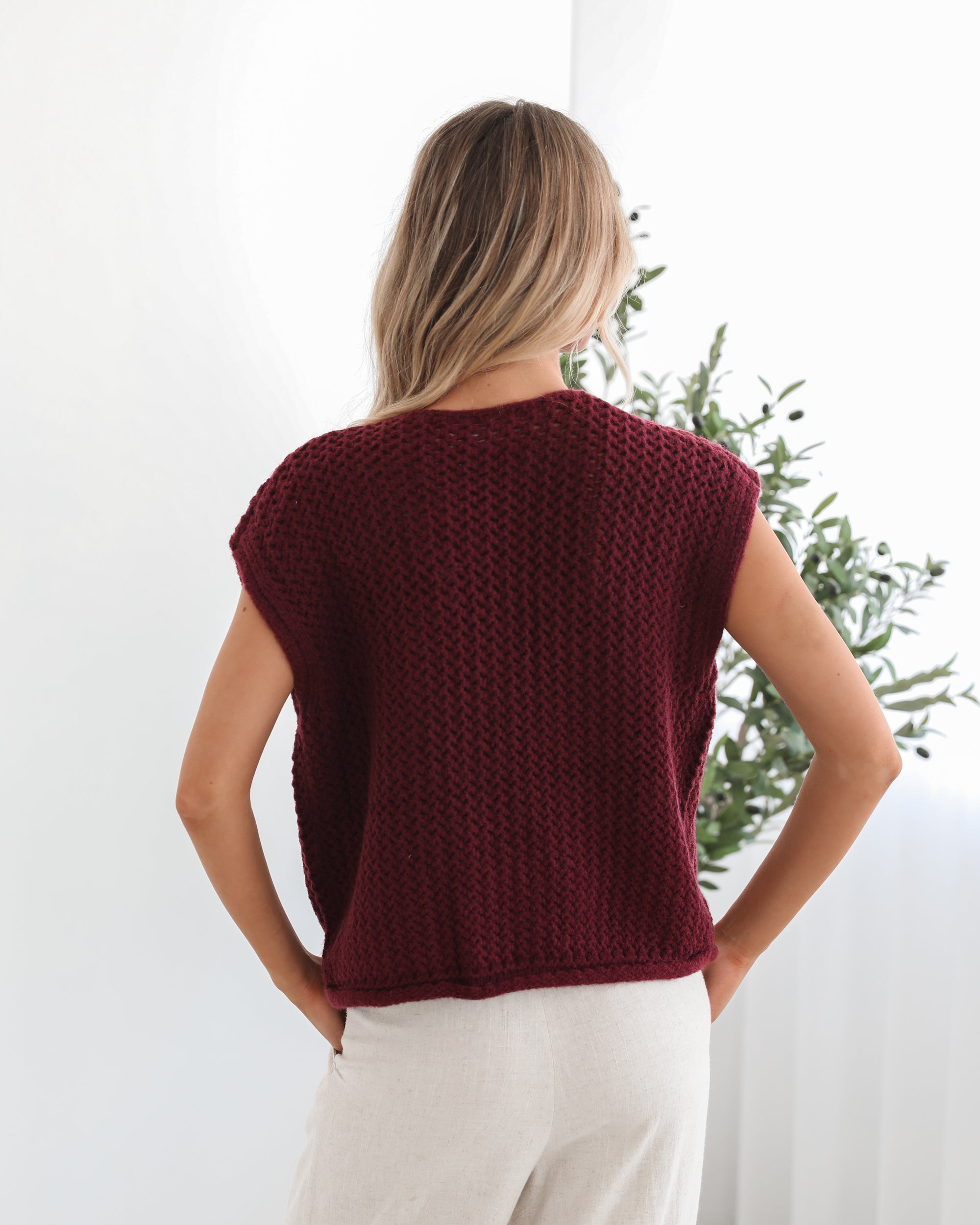 A woman wearing a sleeveless wine-colored knit top with round neckline and center-front buttons, paired with light-colored pants.