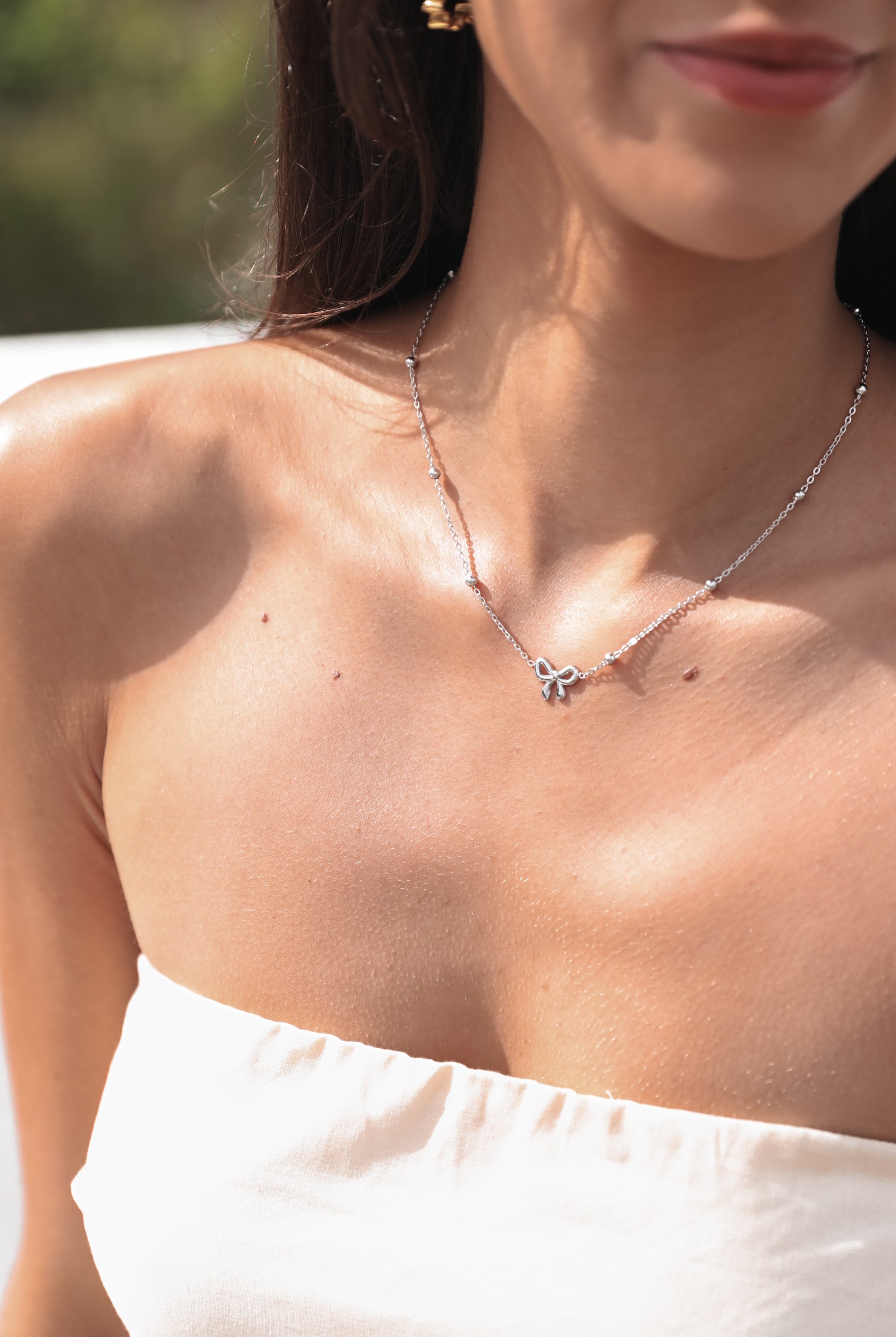 A close-up image of a woman's neckline, showcasing a silver-colored necklace with a chain and bow-shaped pendant.
