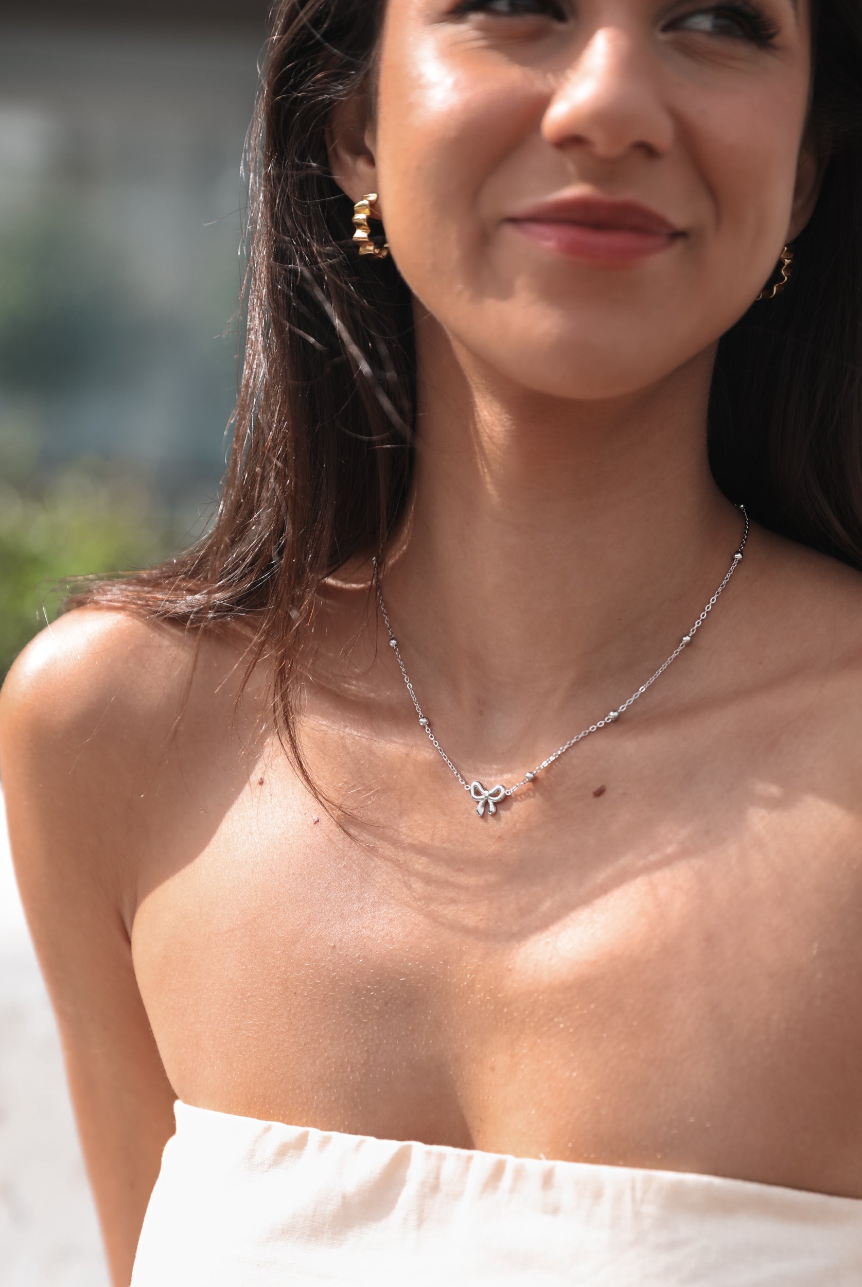 A close-up image of a woman's neckline, showcasing a silver-colored necklace with a chain and bow-shaped pendant.