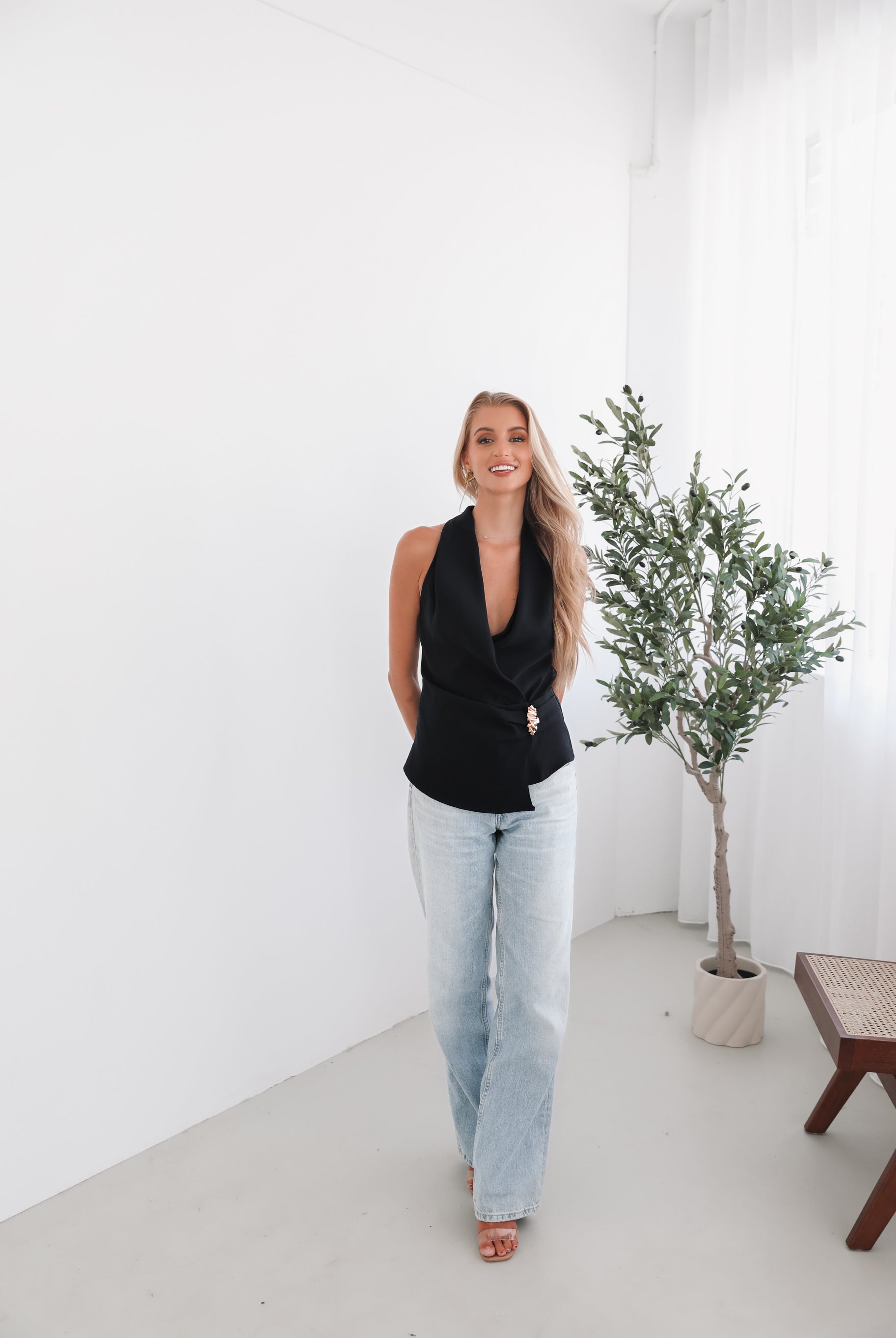 A woman wearing a navy sleeveless top with a plunge neckline and overlap bodice, paired with light jeans. She has blonde hair and is smiling.