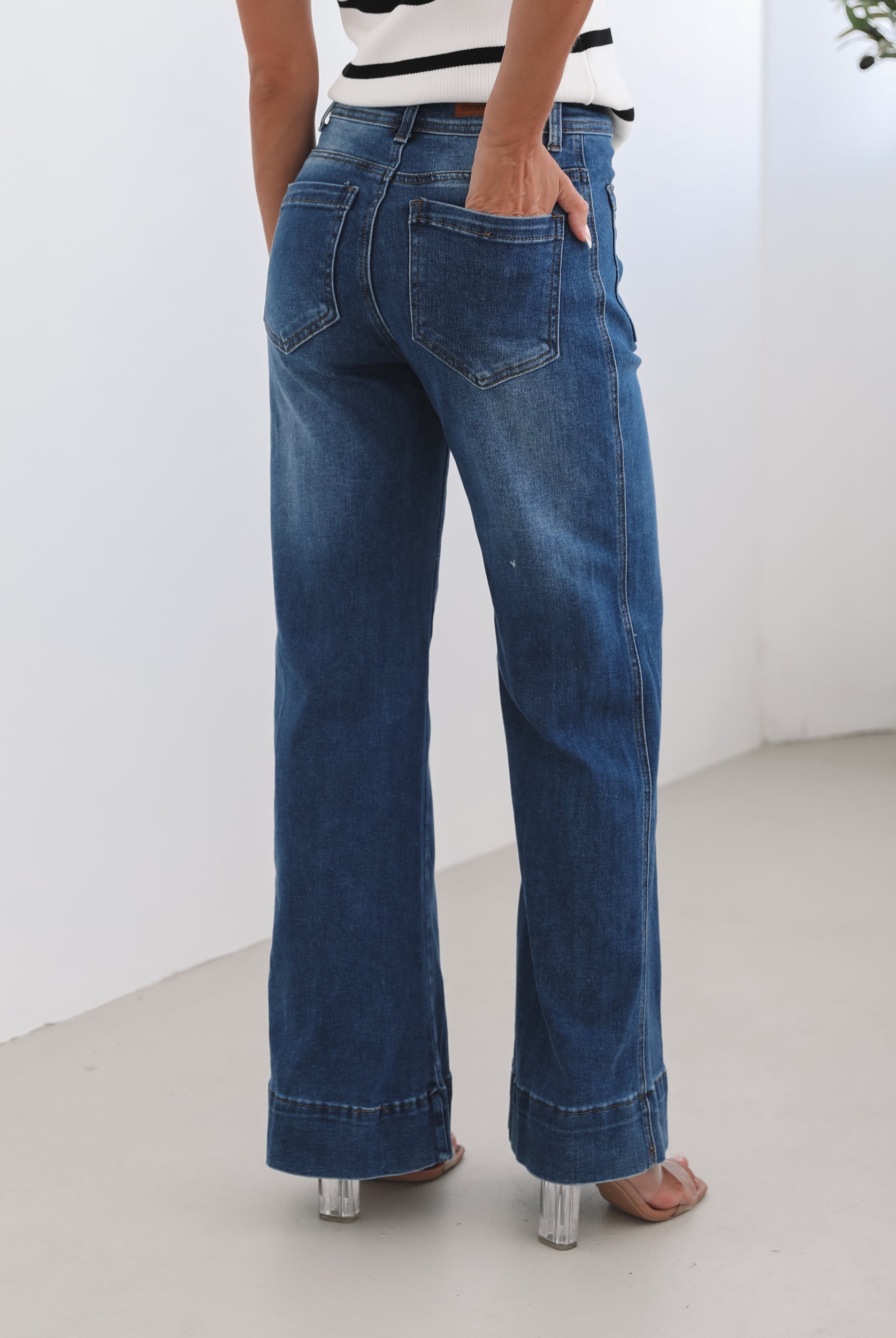 A woman standing in a room with light decor, wearing a striped top and mid-blue Yzabel Jeans with a high waist fit and wide leg silhouette.