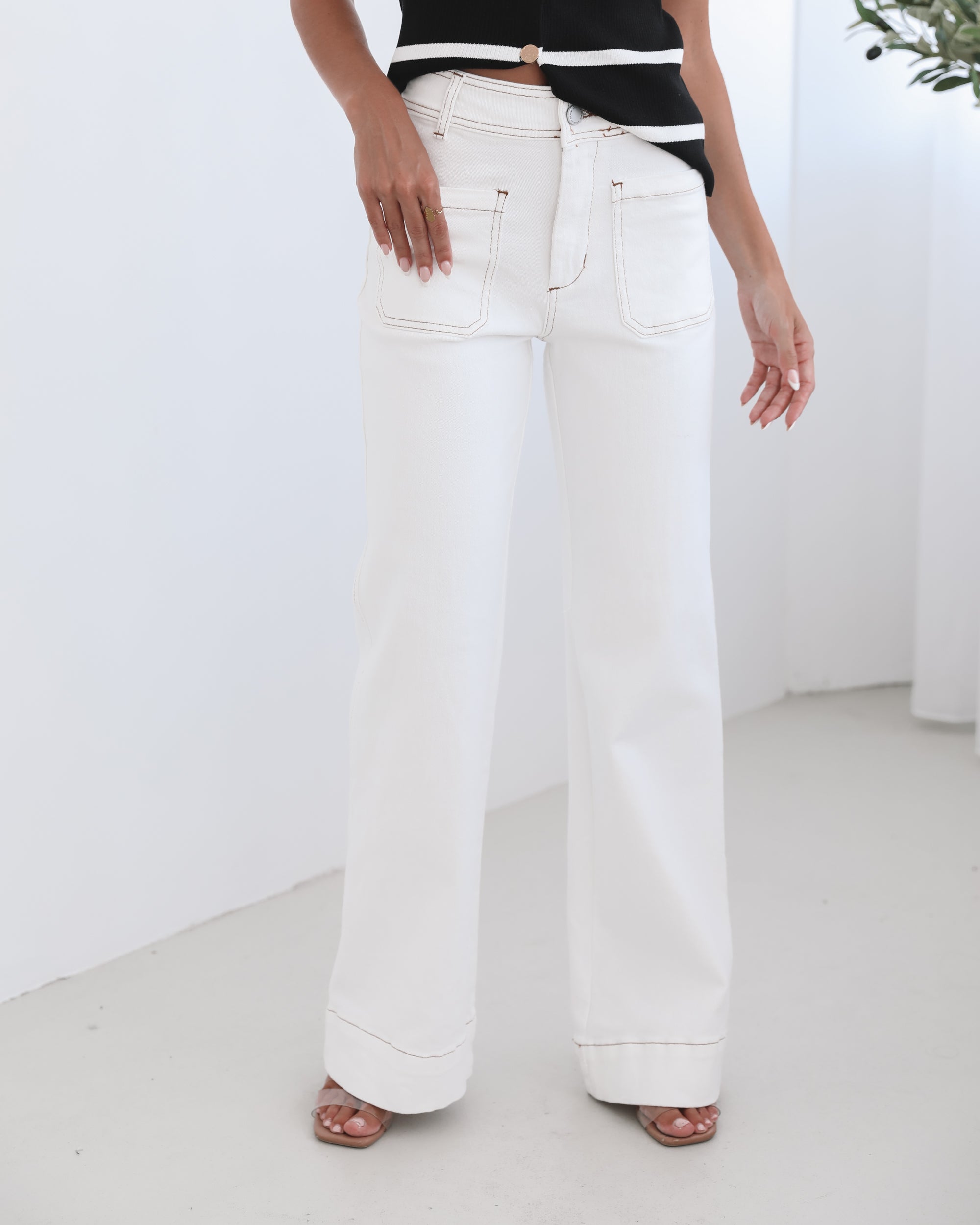A female model wearing white high-waisted jeans with brown contrasting stitches, paired with a striped black and white top.
