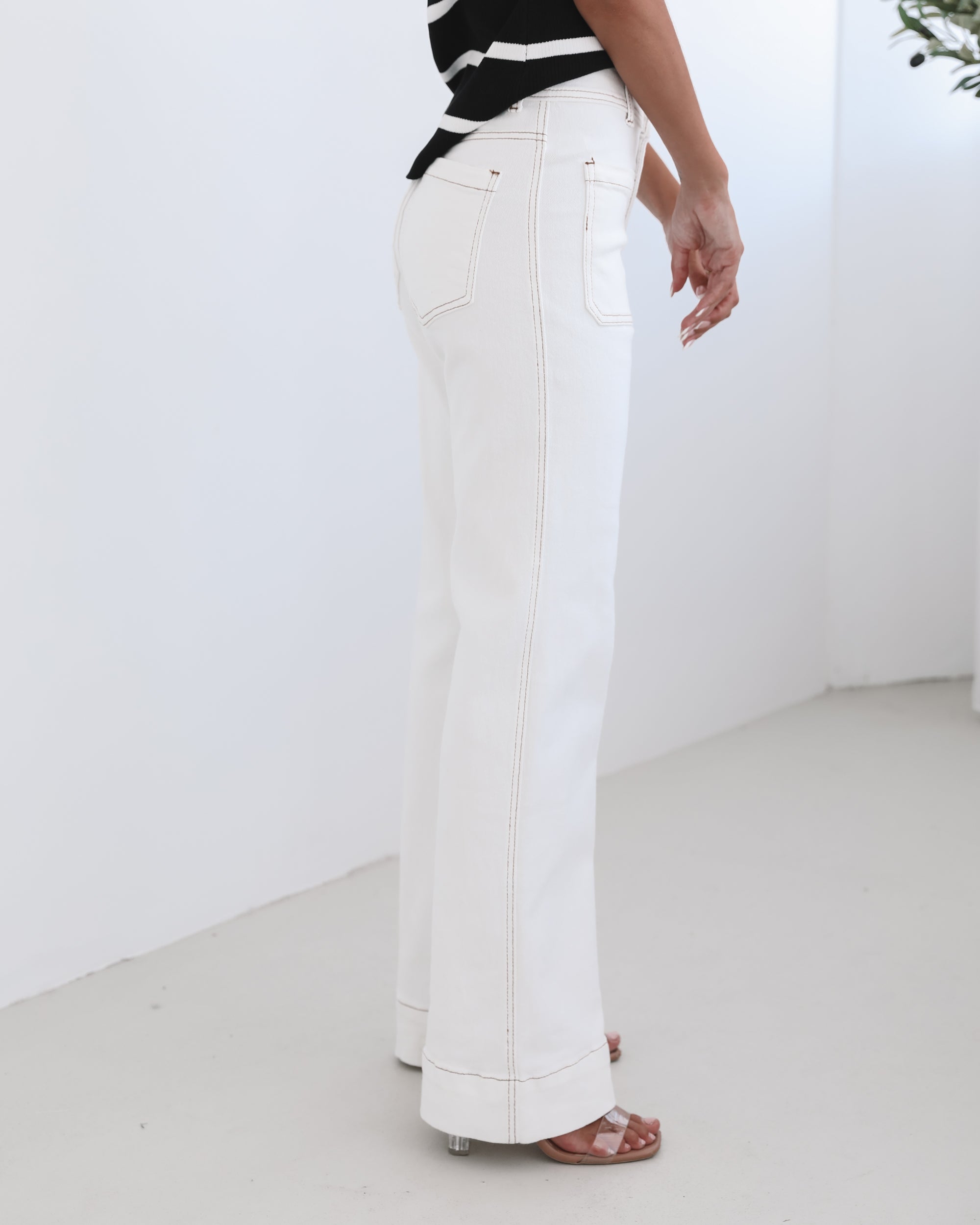 A female model wearing white high-waisted jeans with brown contrasting stitches, paired with a striped black and white top.