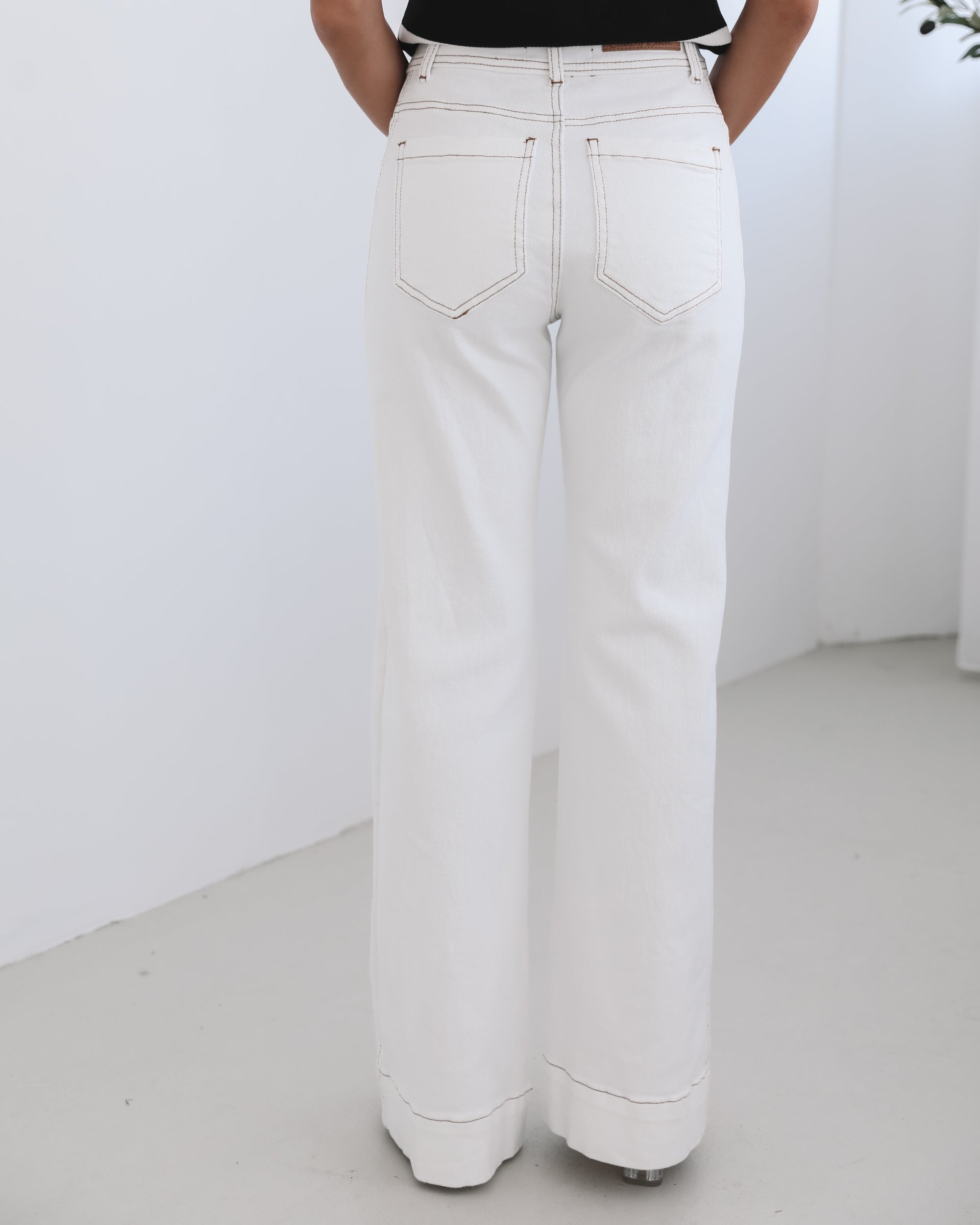 A female model wearing white high-waisted jeans with brown contrasting stitches, paired with a striped black and white top.