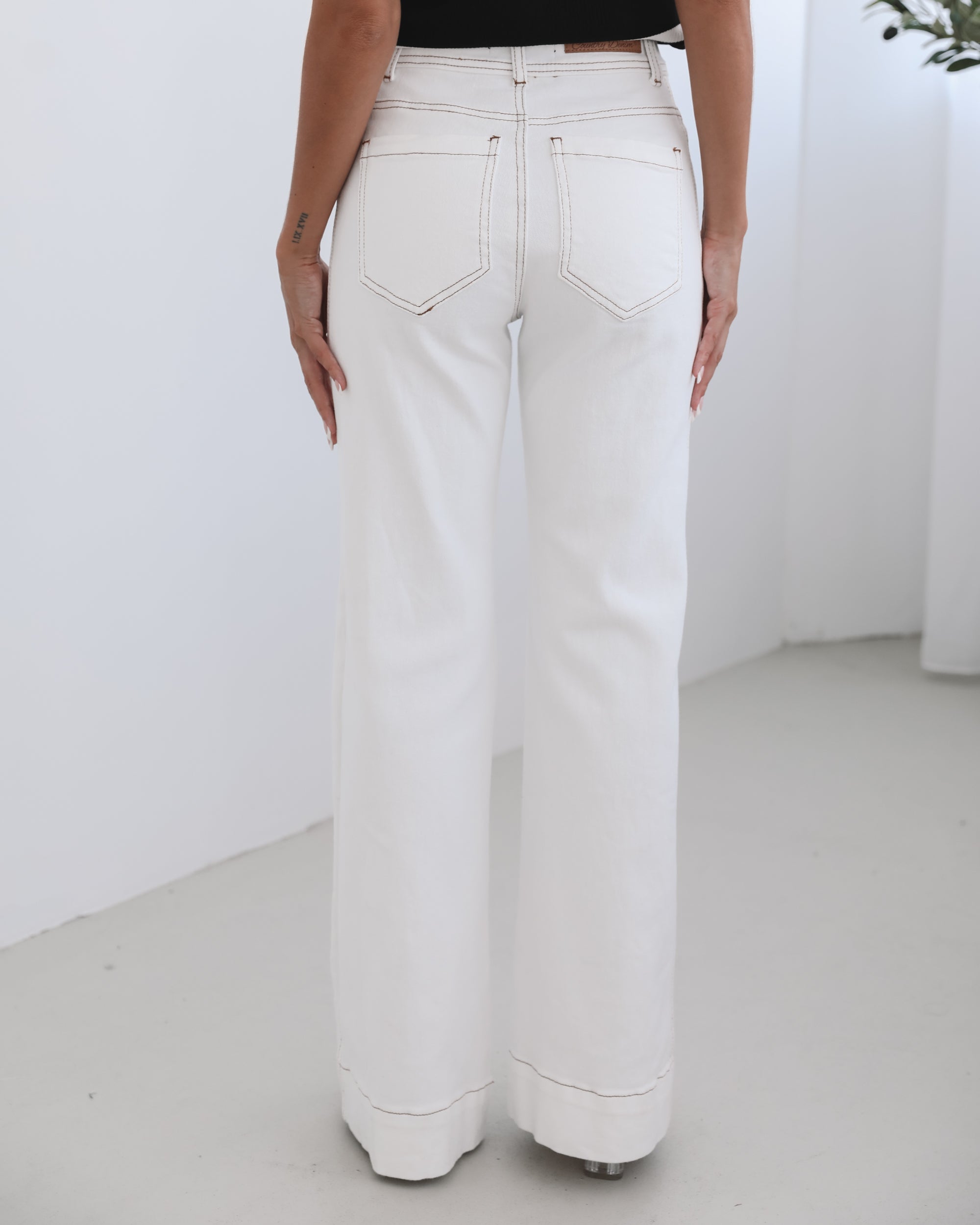 A female model wearing white high-waisted jeans with brown contrasting stitches, paired with a striped black and white top.