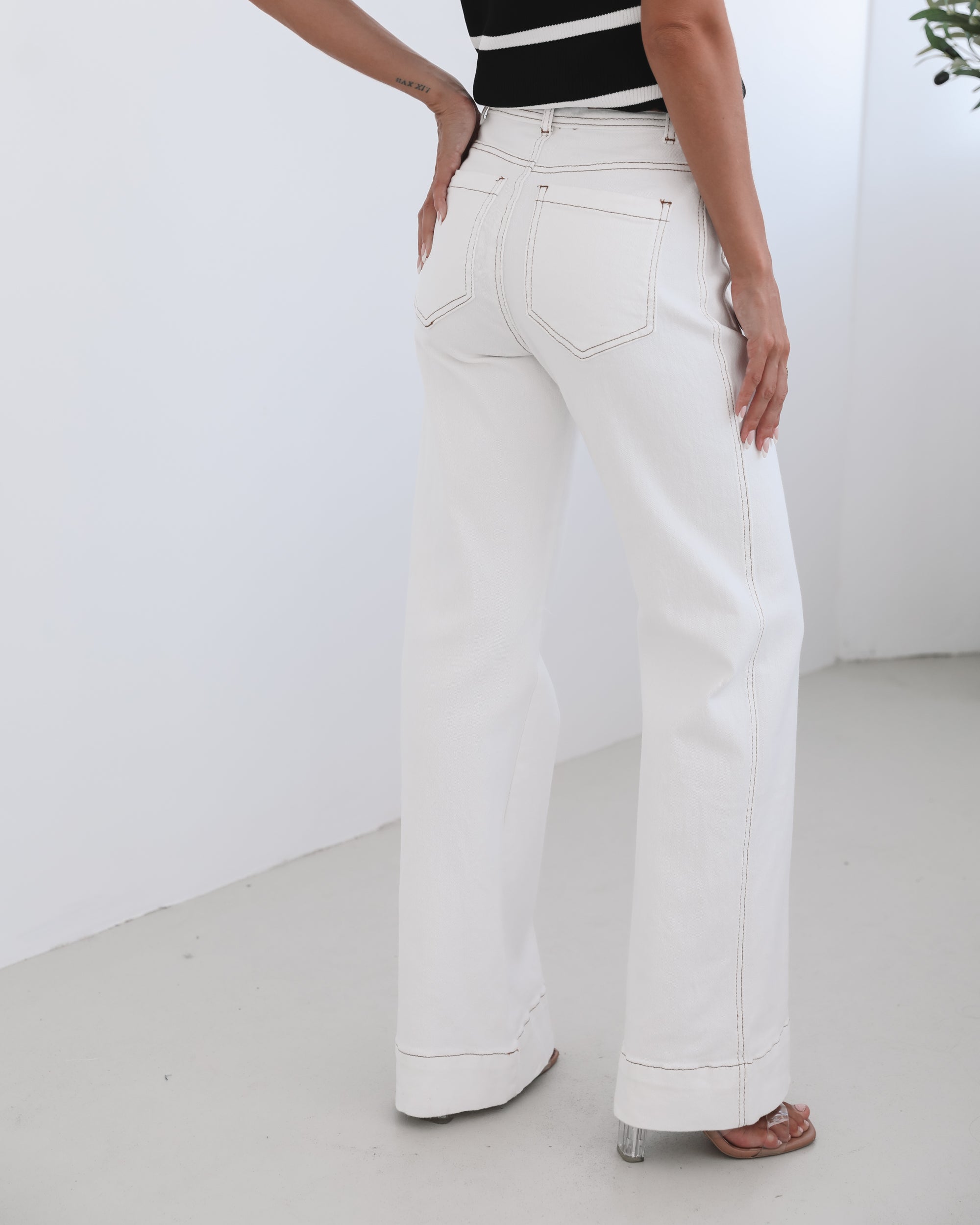 A female model wearing white high-waisted jeans with brown contrasting stitches, paired with a striped black and white top.