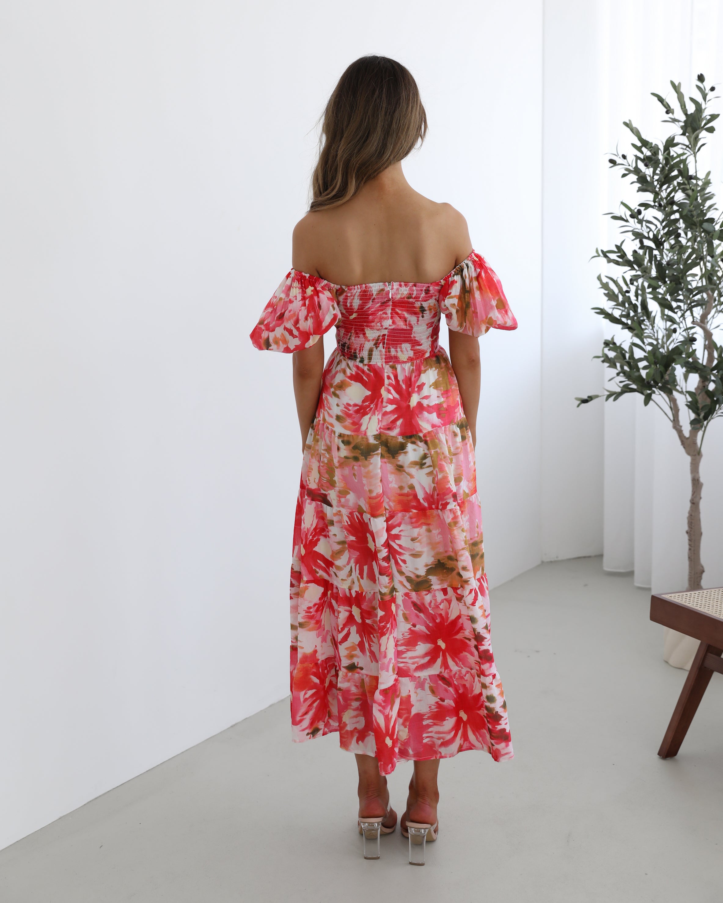 A woman standing next to a chair, wearing a floral print maxi dress with a sweetheart neckline and short balloon sleeves. She is posing in a room with a large window and a plant in the background.
