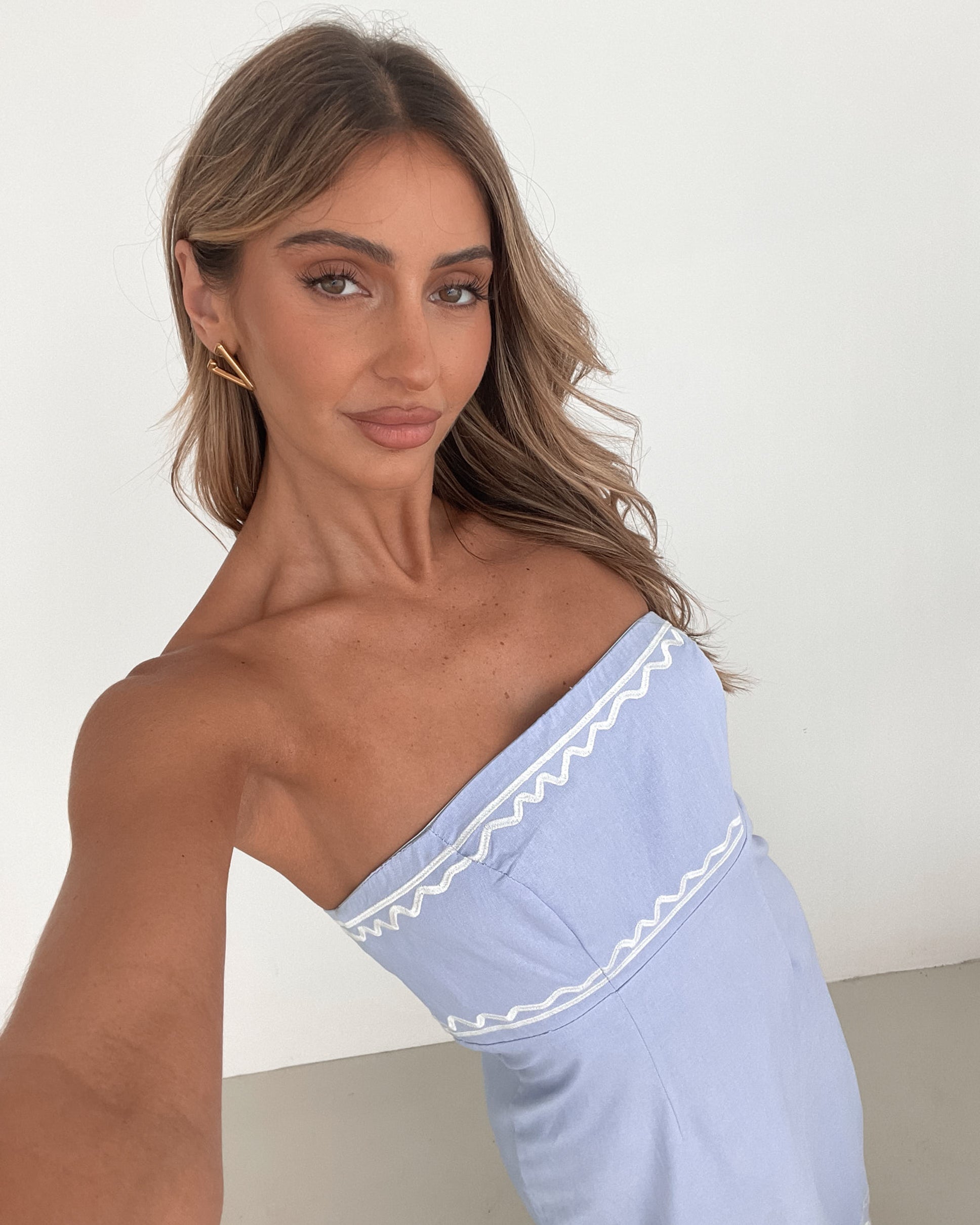 A woman wearing a blue and white floral print maxi dress with a tube style top and fit-to-flare silhouette. She is standing in a room with a white wall and a wooden bench, posing with one hand on her hip.