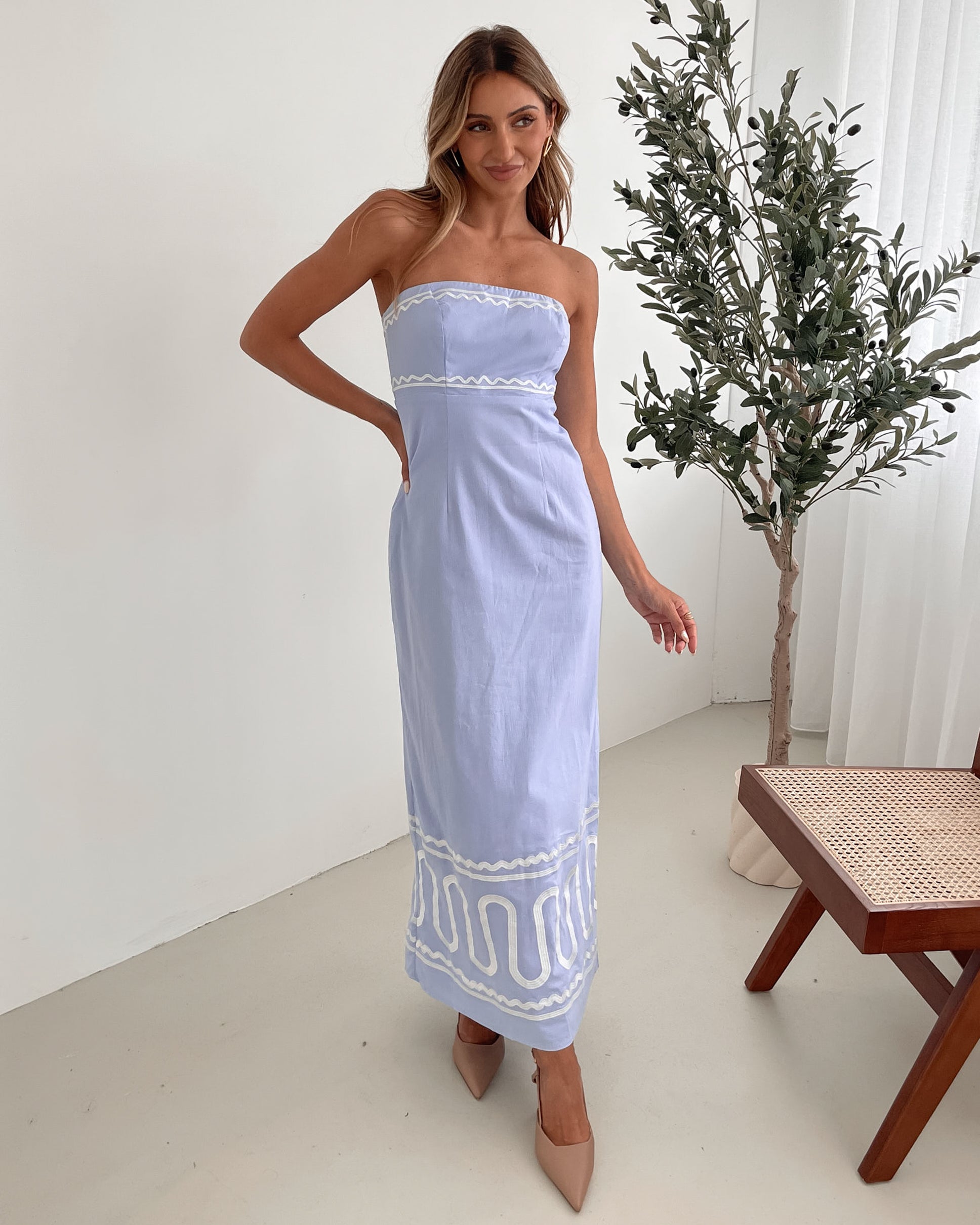 A woman wearing a blue and white floral print maxi dress with a tube style top and fit-to-flare silhouette. She is standing in a room with a white wall and a wooden bench, posing with one hand on her hip.