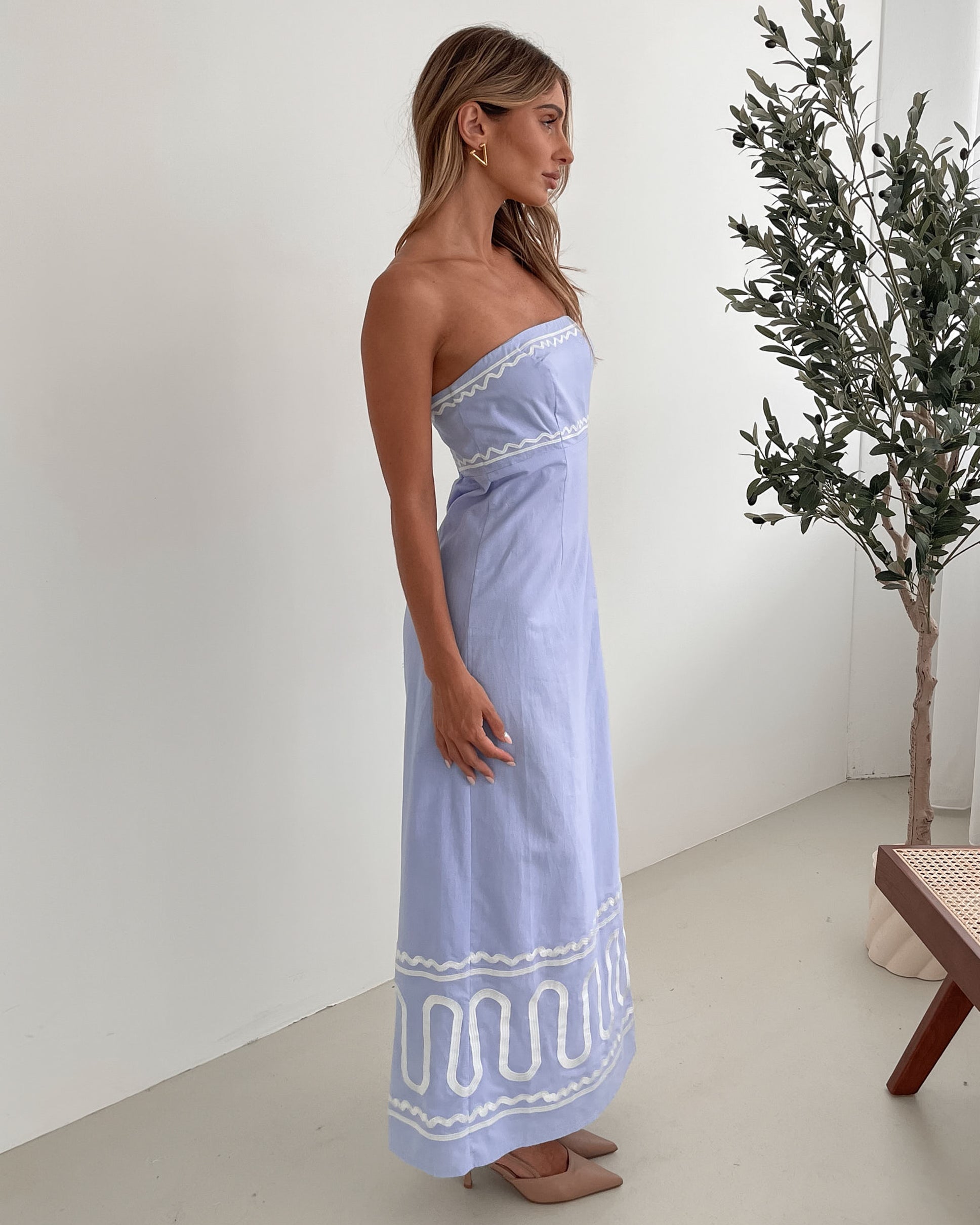 A woman wearing a blue and white floral print maxi dress with a tube style top and fit-to-flare silhouette. She is standing in a room with a white wall and a wooden bench, posing with one hand on her hip.