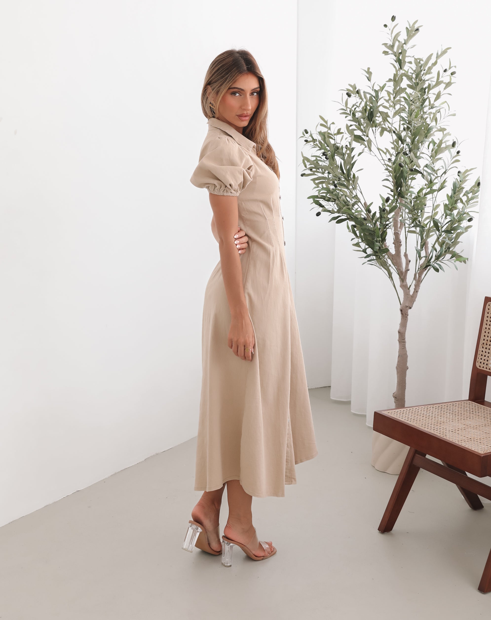 A woman standing in a room with a beige dress with a shirt collar, short puff sleeves, and a-line skirt with a midaxi length hem.