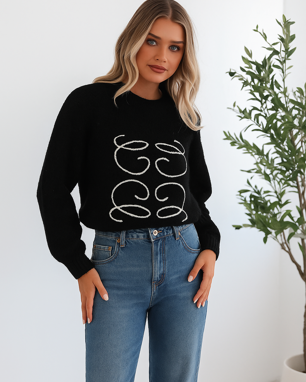 A woman with long, wavy blonde hair is standing indoors near a potted plant, wearing a black sweater with a white abstract design on the front and high-waisted blue jeans. She is looking at the camera with a neutral expression and has one hand resting on her hip. The background is minimalistic with white walls.