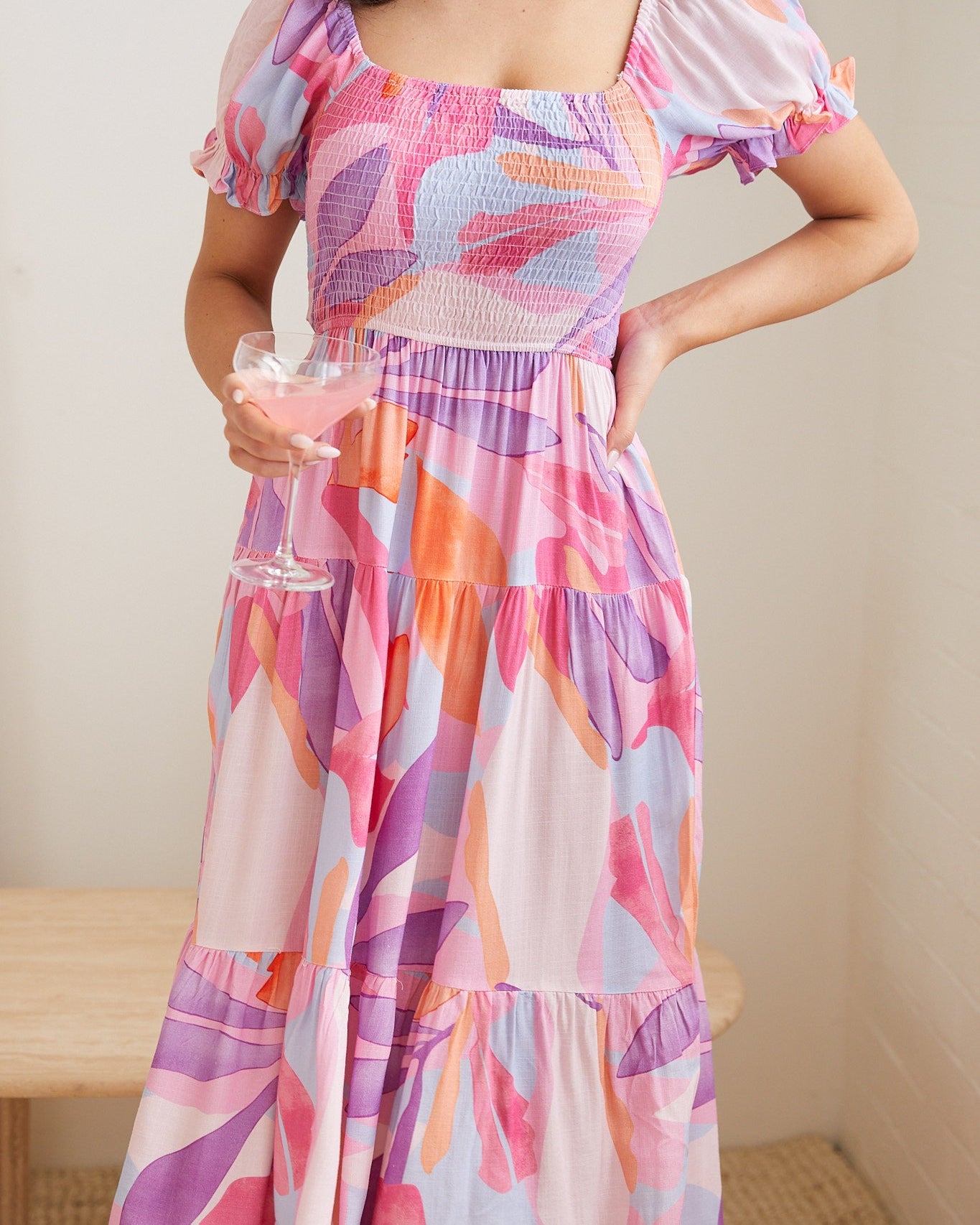 A woman wearing a colorful, abstract-patterned midi dress in shades of pink, purple, and orange. The dress features puff sleeves, a smocked bodice, and a tiered skirt. She poses indoors near a modern wooden table with a white vase holding white flowers, and a large arched mirror in the background.