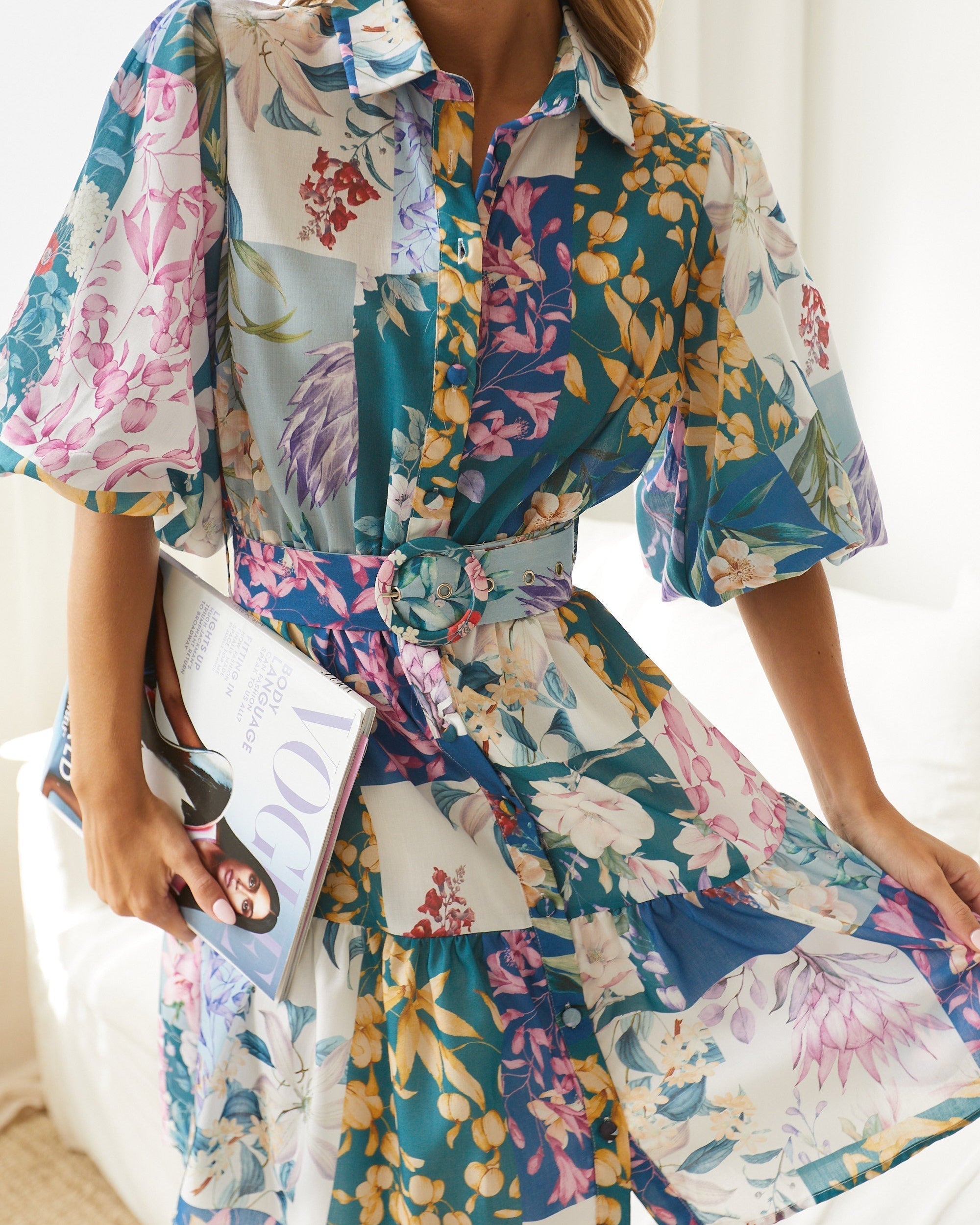 A woman wearing a short, multicolored floral dress with puff sleeves and a belted waist. The dress features a patchwork design with vibrant flower patterns in shades of pink, blue, yellow, and green. She is standing indoors in front of a white couch, looking to the side with a gentle smile.