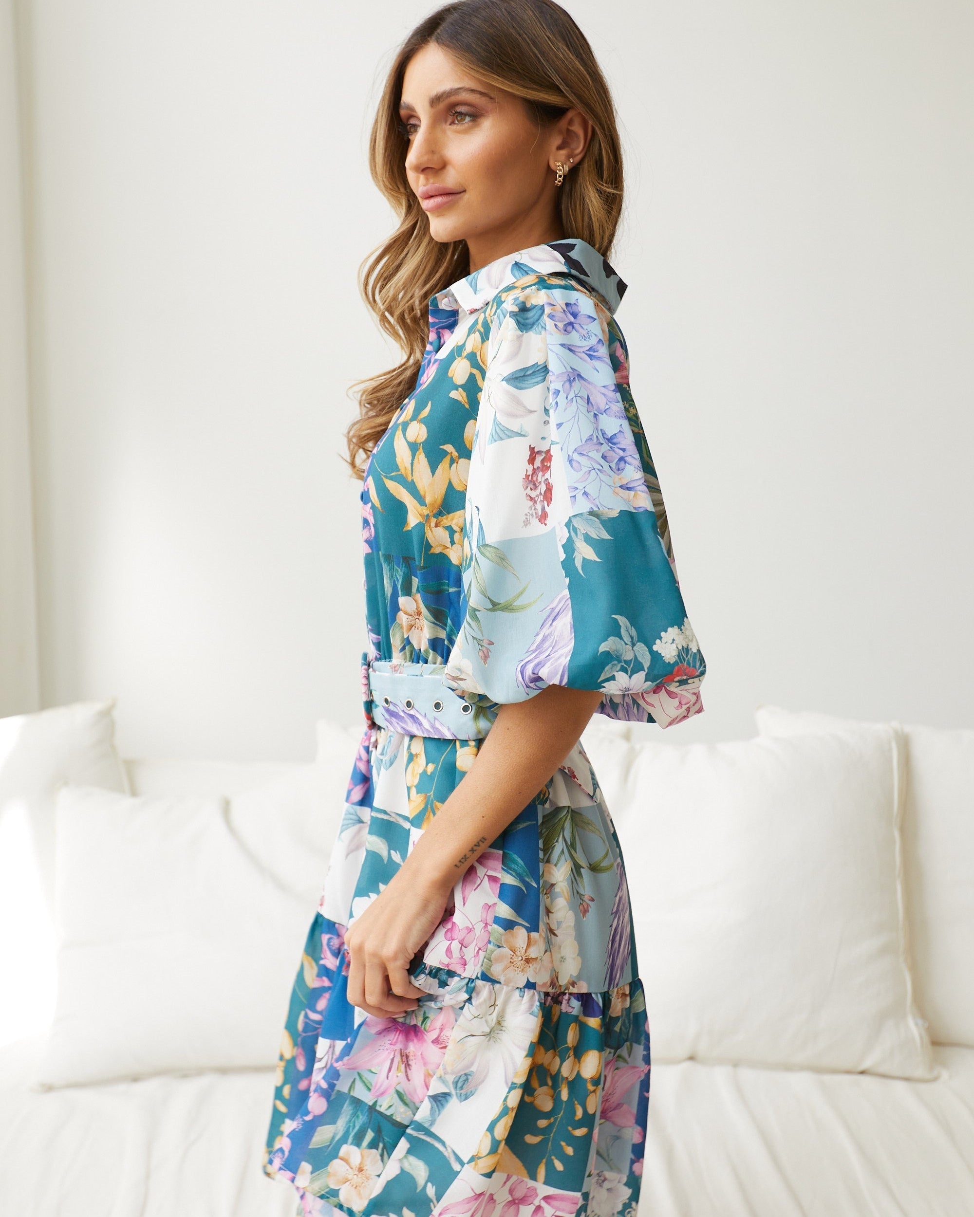 A woman wearing a short, multicolored floral dress with puff sleeves and a belted waist. The dress features a patchwork design with vibrant flower patterns in shades of pink, blue, yellow, and green. She is standing indoors in front of a white couch, looking to the side with a gentle smile.