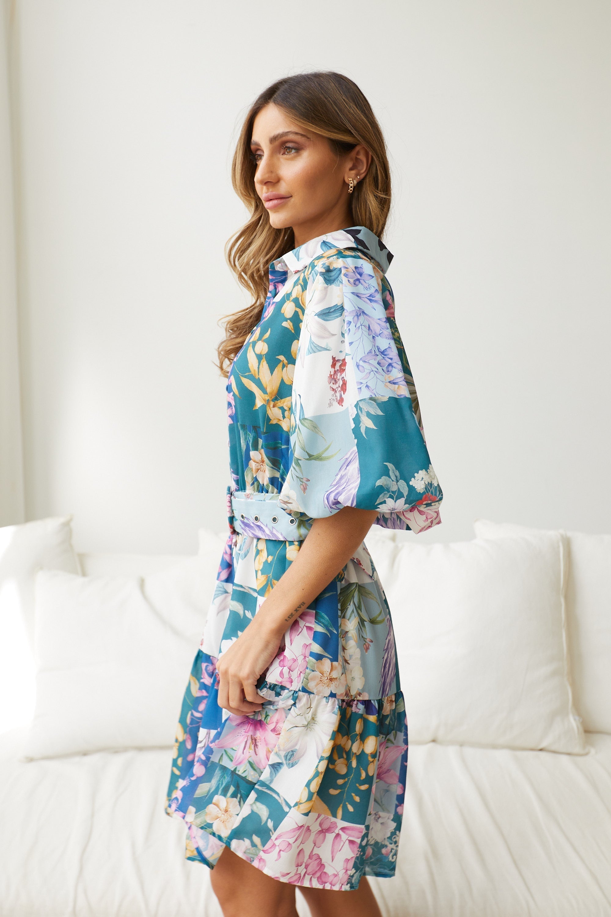 A woman wearing a short, multicolored floral dress with puff sleeves and a belted waist. The dress features a patchwork design with vibrant flower patterns in shades of pink, blue, yellow, and green. She is standing indoors in front of a white couch, looking to the side with a gentle smile.