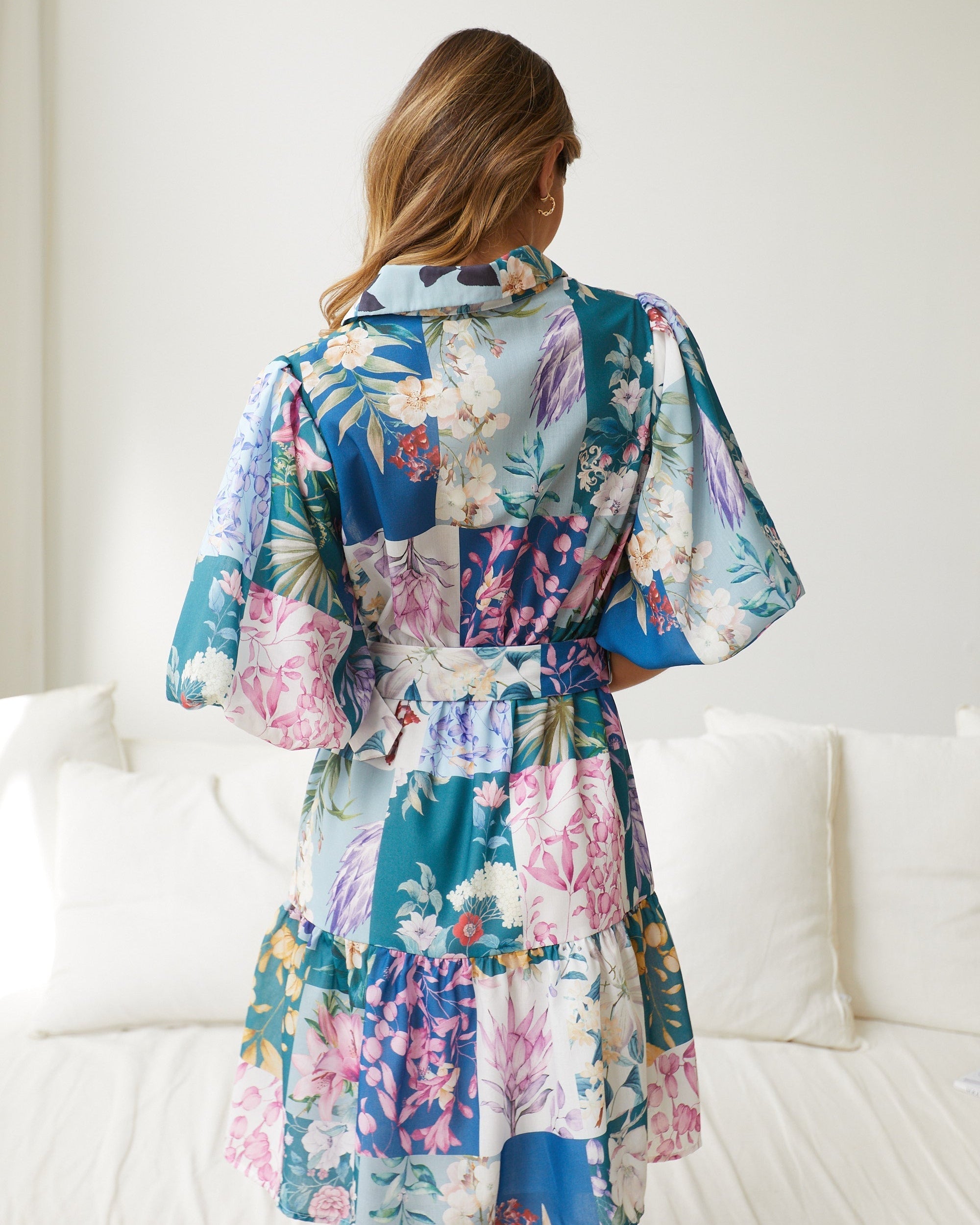 A woman wearing a short, multicolored floral dress with puff sleeves and a belted waist. The dress features a patchwork design with vibrant flower patterns in shades of pink, blue, yellow, and green. She is standing indoors in front of a white couch, looking to the side with a gentle smile.