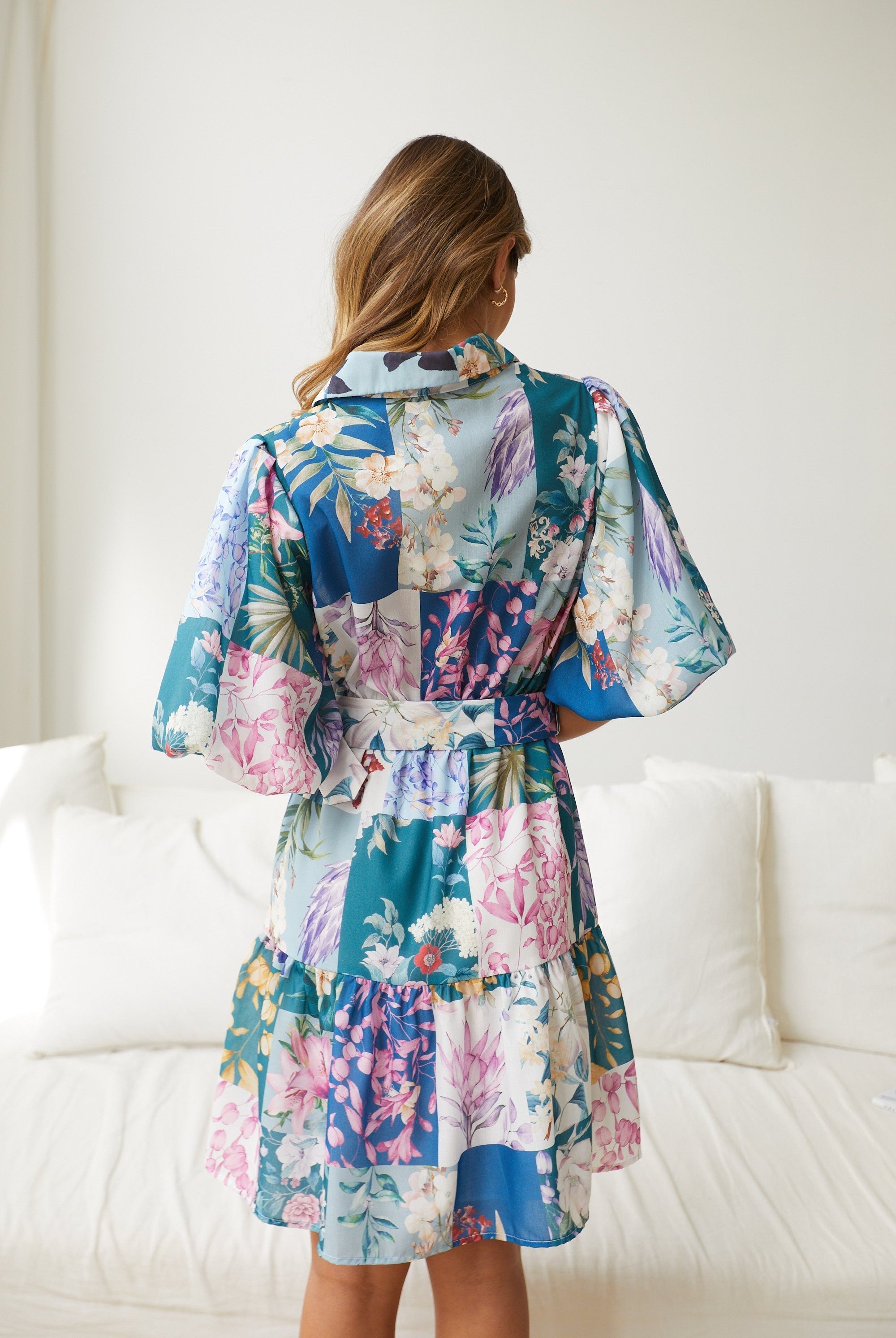 A woman wearing a short, multicolored floral dress with puff sleeves and a belted waist. The dress features a patchwork design with vibrant flower patterns in shades of pink, blue, yellow, and green. She is standing indoors in front of a white couch, looking to the side with a gentle smile.